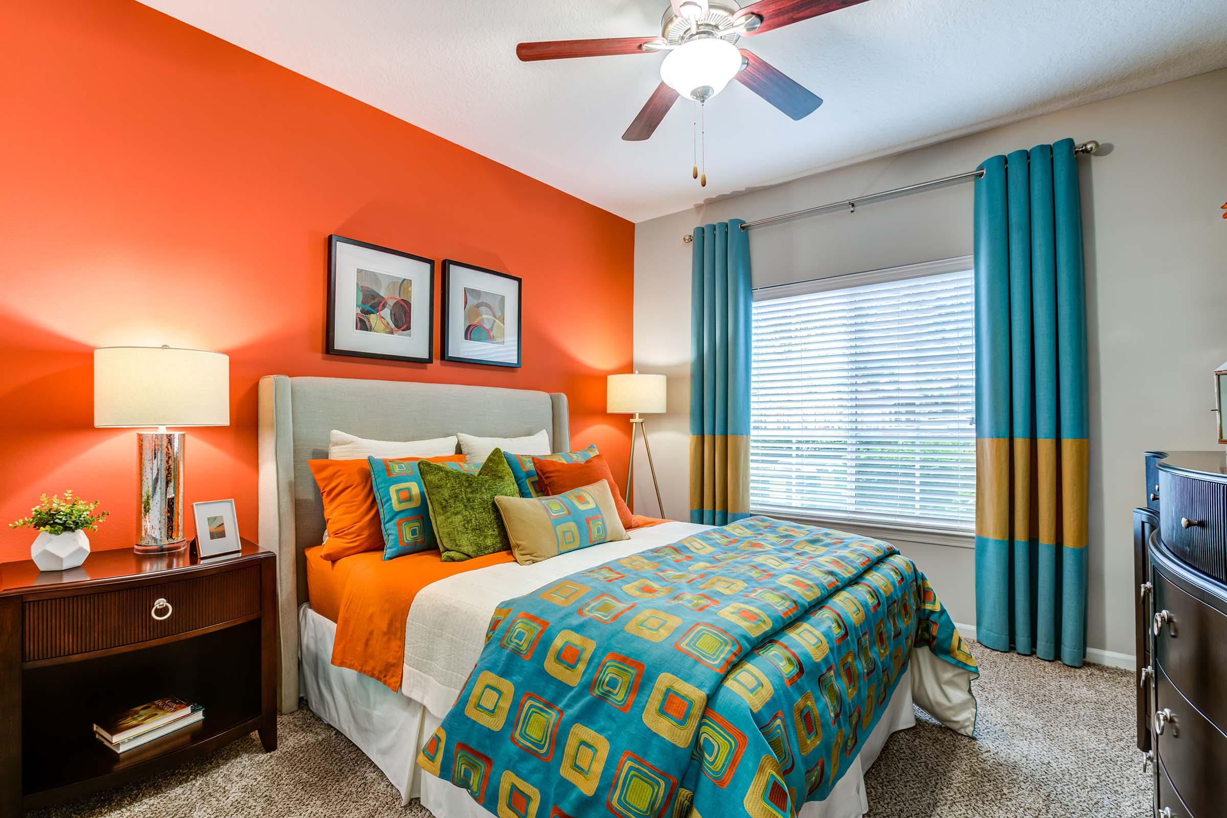 A cozy bedroom featuring an orange accent wall and bright bedding with geometric patterns. The room includes a comfortable bed with colorful pillows, a nightstand with a lamp, and a stylish dresser. Large windows with turquoise curtains allow natural light in, creating a warm and inviting atmosphere.