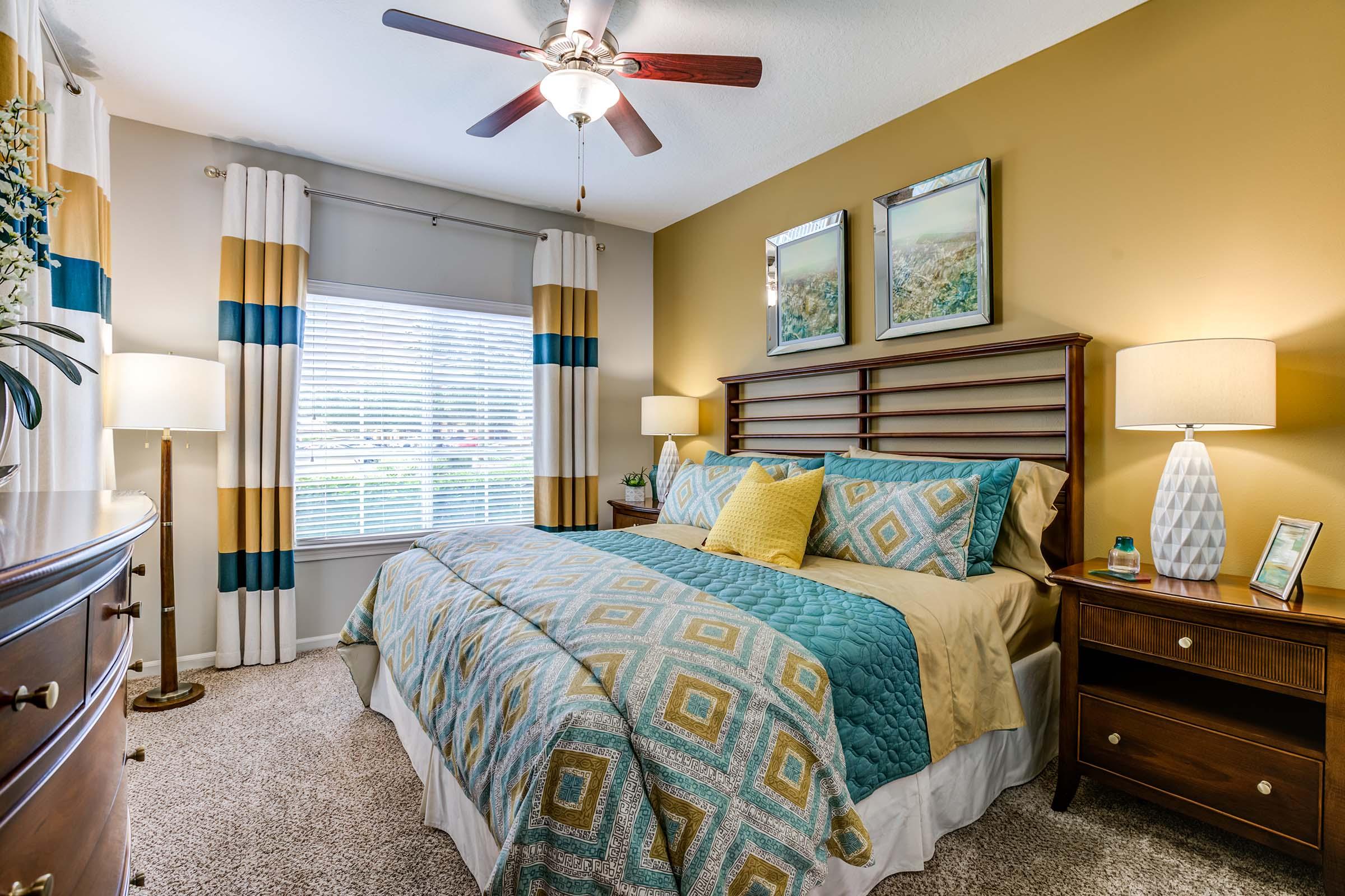 A cozy bedroom featuring a wooden bed with a blue and yellow patterned comforter, two decorative pillows, and matching curtains. There are bedside lamps on wooden nightstands, a dresser with a mirror, and artwork on the wall. Large windows let in natural light.