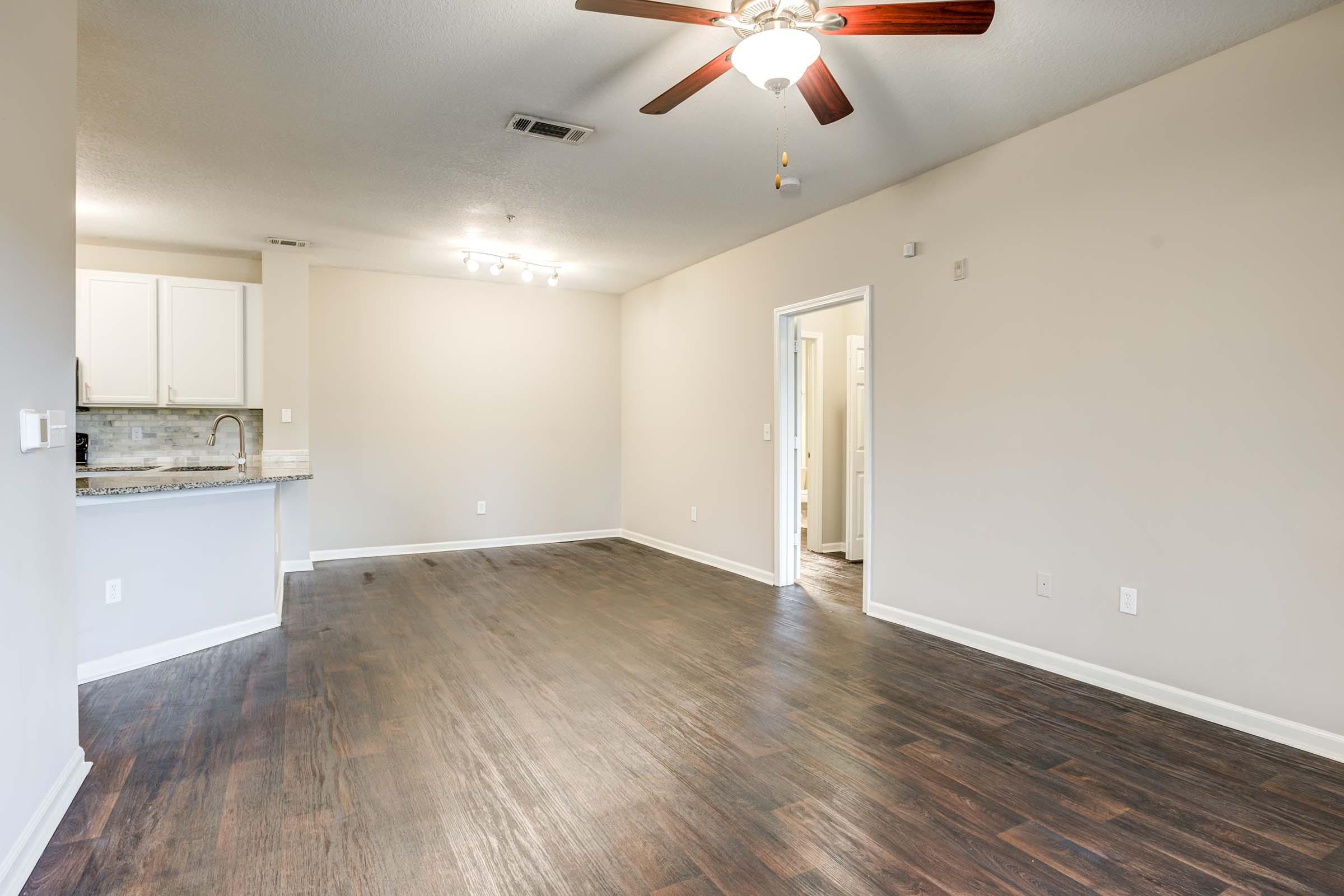 Empty living room featuring hardwood floors, a ceiling fan, and natural light. The space includes a kitchen area with light-colored cabinets and a doorway leading to a bathroom or other room. The walls are painted a neutral tone, creating a bright and open atmosphere.