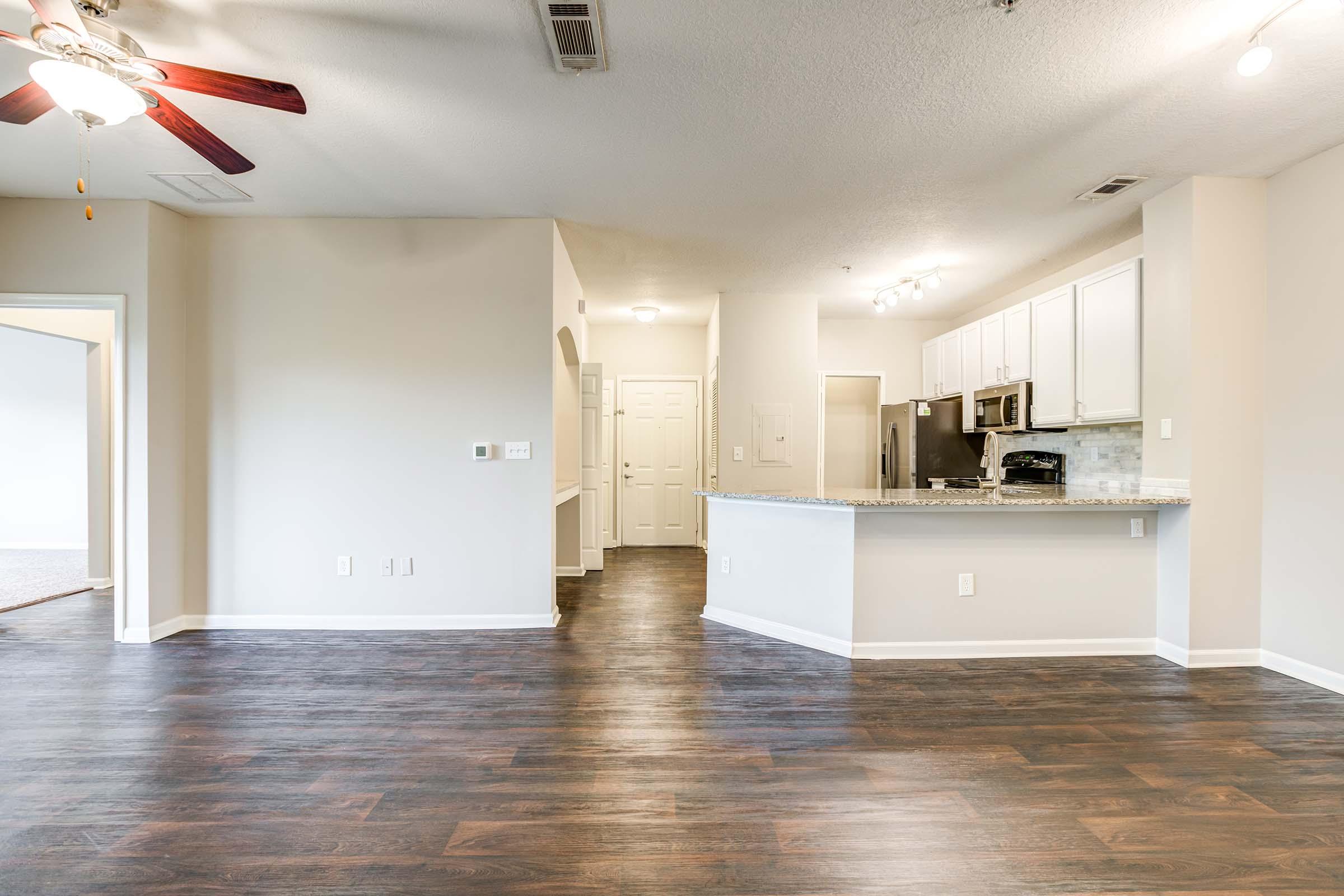 View of an open-concept living space featuring a modern kitchen with white cabinets and stainless steel appliances, a spacious living area with dark wood flooring, and a ceiling fan. The room is well-lit with natural light and has a doorway leading to another room.