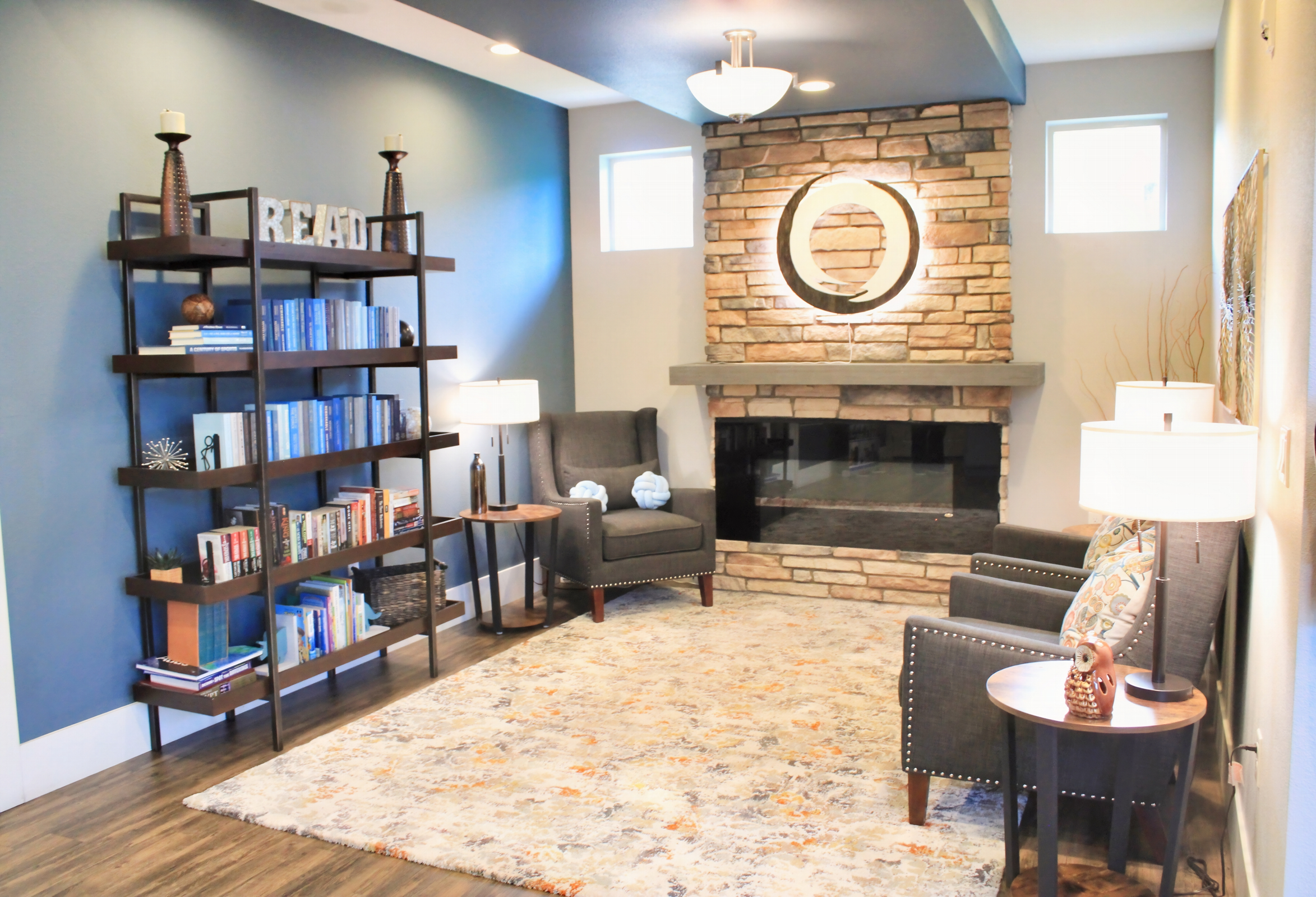 A cozy living room featuring a modern bookshelf with books, two comfortable armchairs, and a round mirror above a stone fireplace. Soft lighting from two side tables creates a warm atmosphere, complemented by an area rug that adds texture to the space.