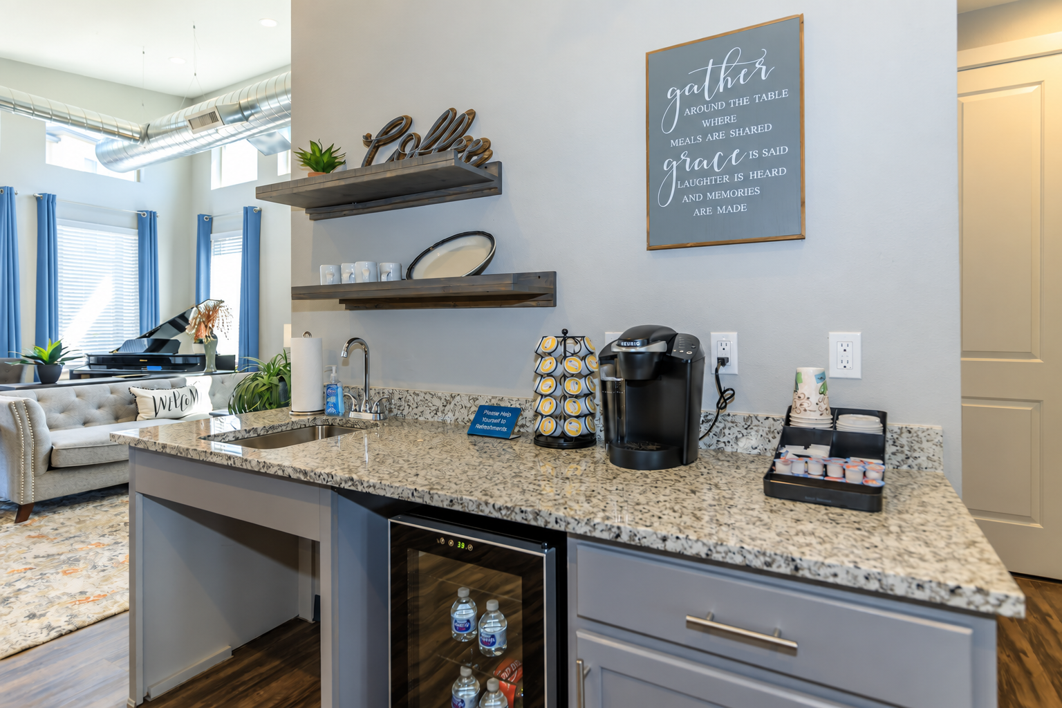 A modern kitchen nook featuring a granite countertop with a small sink, a coffee maker, and a mini-fridge stocked with bottled water. There are decorative shelves above with plates and a wall sign that reads "Gather around the table." The background shows a comfortable living space with a piano and a cozy seating area.
