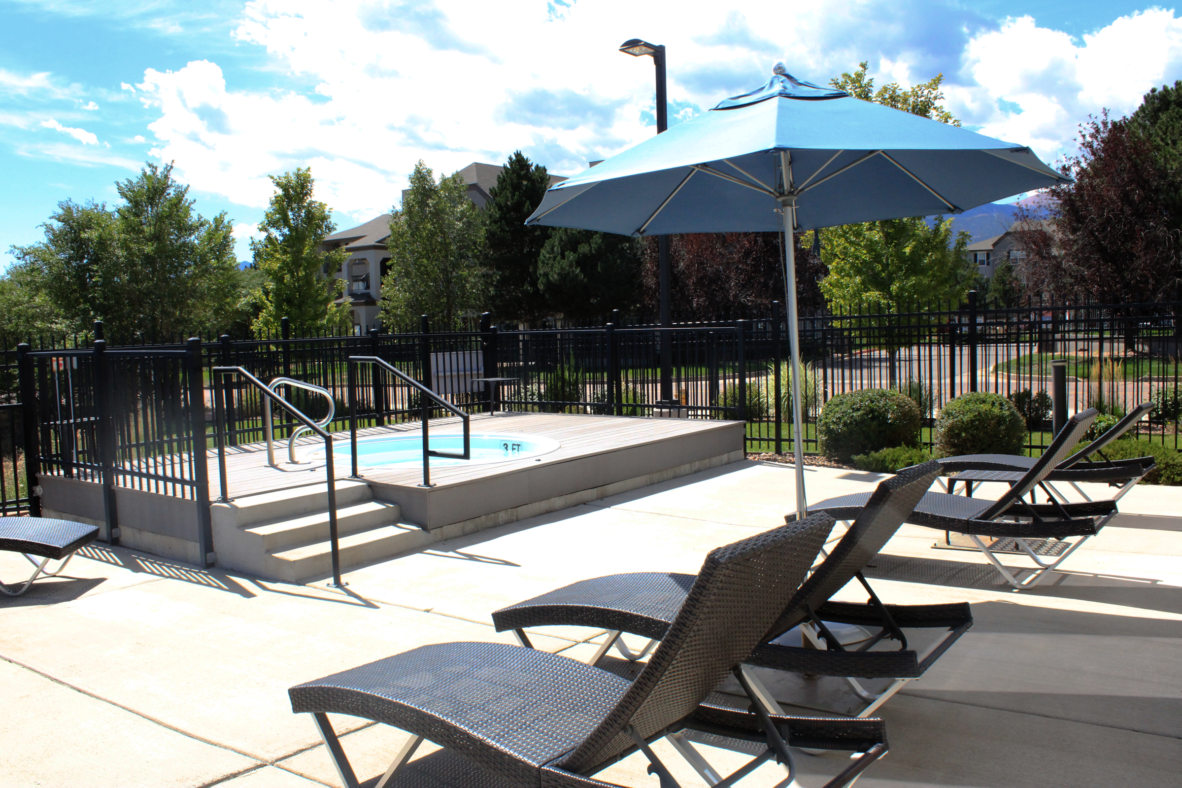 A bright outdoor pool area featuring a small hot tub, surrounded by a secure fence. Several lounge chairs are positioned nearby for relaxation, with a large blue umbrella providing shade. Lush greenery and trees add a natural touch, under a partly cloudy sky.
