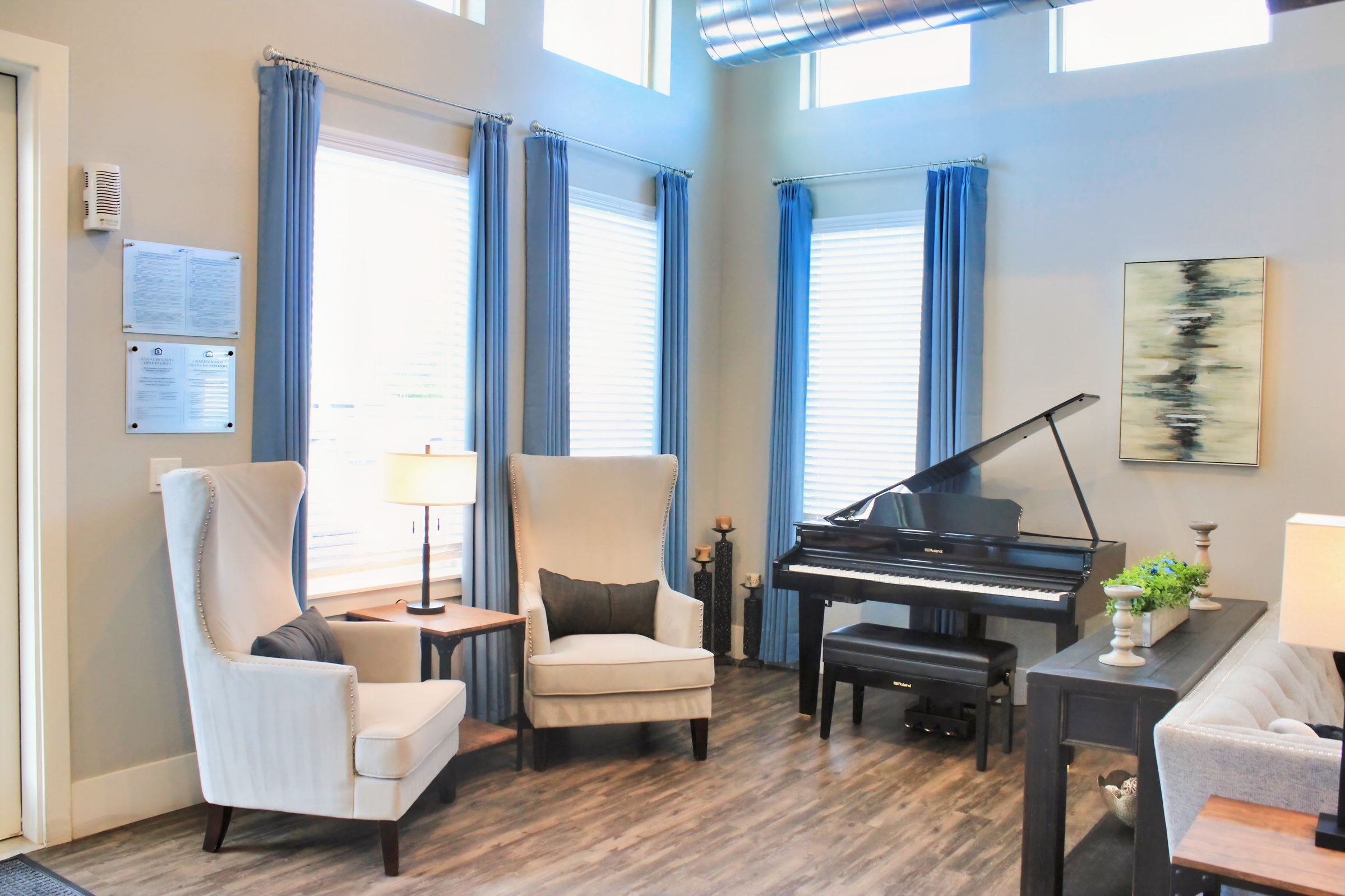 A cozy living room featuring two modern wingback chairs with cushions, a wooden side table with a lamp, and a black grand piano. Large windows with blue curtains allow natural light to fill the space. A piece of abstract art is displayed on the wall, and the flooring is a warm, wood-like finish.