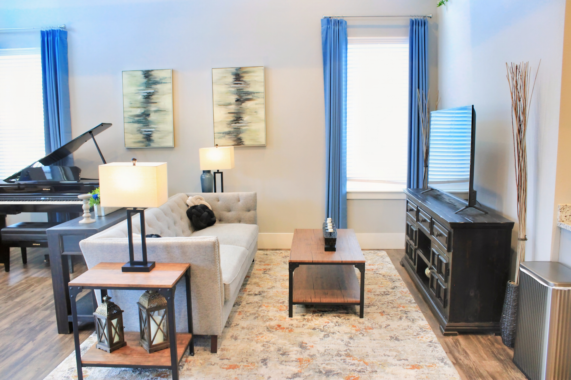 A modern living room featuring a light gray sofa with a decorative throw, a wooden coffee table, and a dark wooden TV stand. Two lamps provide soft lighting, and there are two blue curtains framing a window. Wall art consisting of abstract paintings complements the decor, along with a piano in the background.