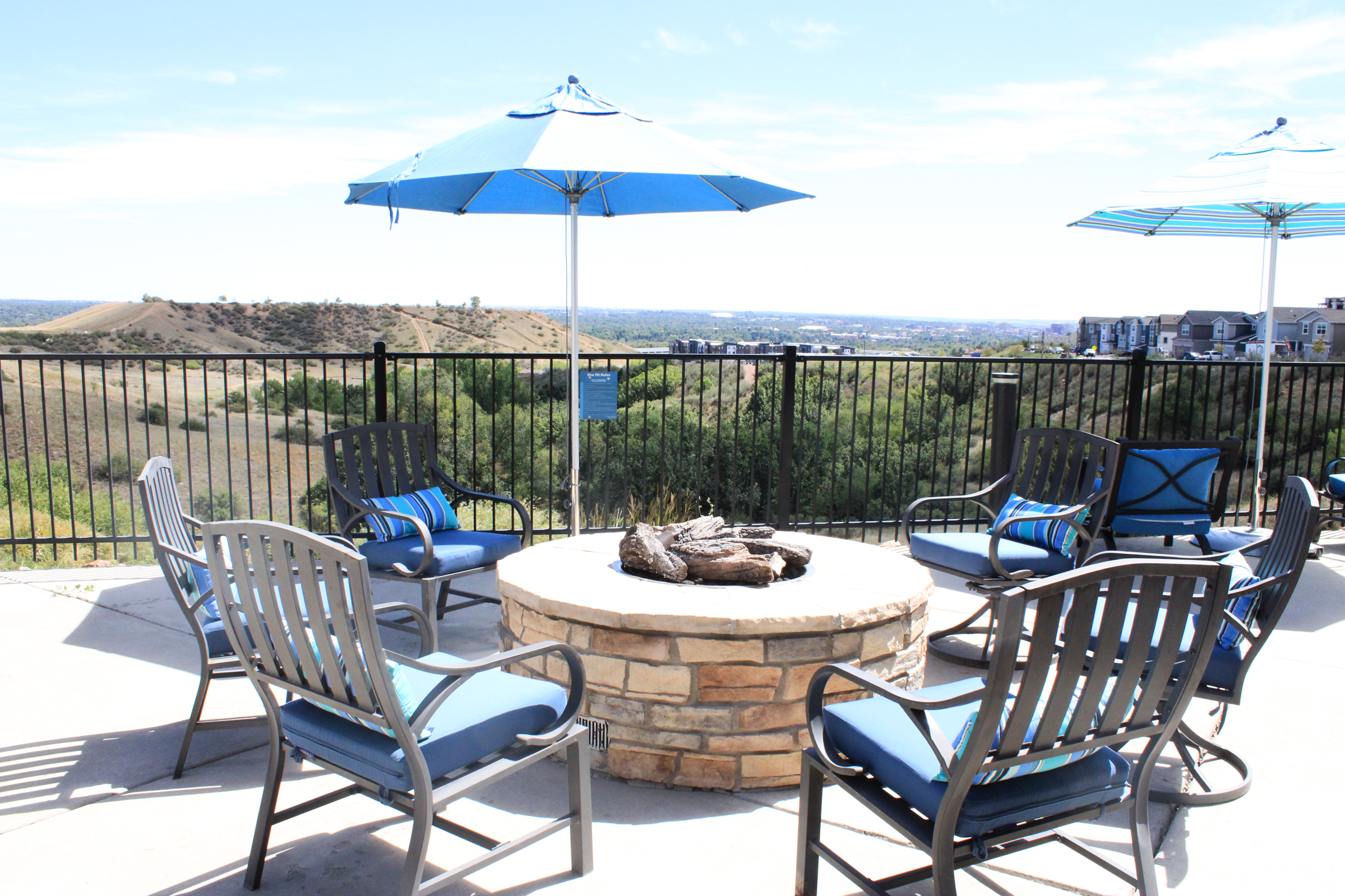 A cozy outdoor seating area with a circular stone fire pit surrounded by metal chairs with blue cushions. Two blue umbrellas provide shade, and the backdrop features rolling hills and a view of a nearby settlement under a clear sky.