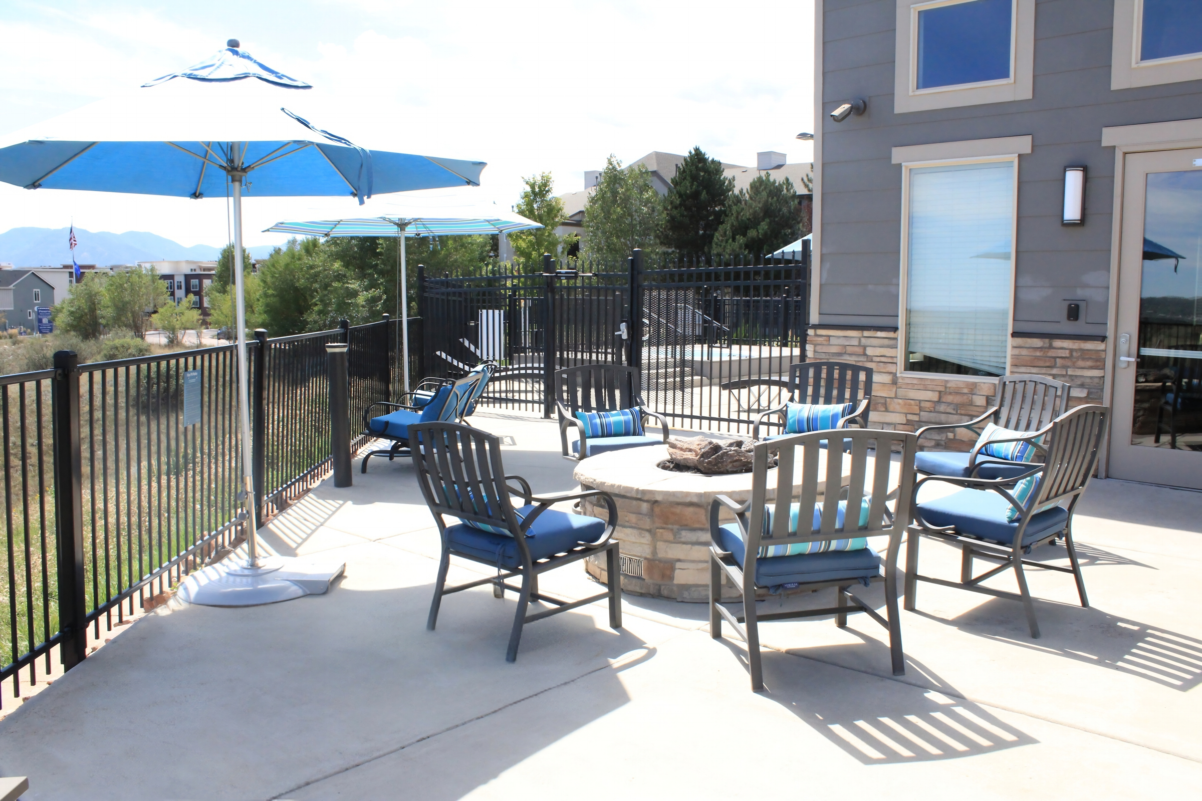 A patio area featuring a circular stone fire pit surrounded by several blue cushioned chairs. To one side, a large blue umbrella provides shade. The space is enclosed by a black fence, with greenery and buildings visible in the background under a clear sky.