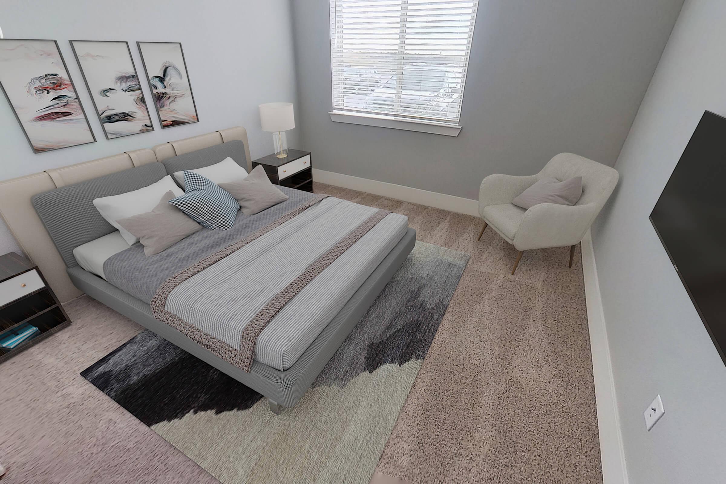 A cozy bedroom featuring a modern bed with gray and white bedding, accent pillows, and a plush area rug. There are framed artworks on the walls, a bedside lamp, a small nightstand, a comfortable armchair in the corner, and a wall-mounted television opposite the bed, with natural light coming through a window.