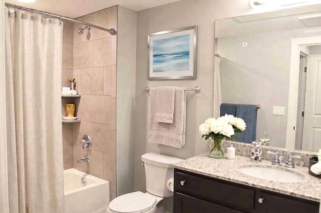 A modern bathroom featuring beige tiled walls, a shower with a curtain, a white bathtub, and a white toilet. There's a granite countertop with a sink, a vase of white flowers, and neatly arranged towels. A framed picture of a seascape hangs on the wall, adding a touch of decor.