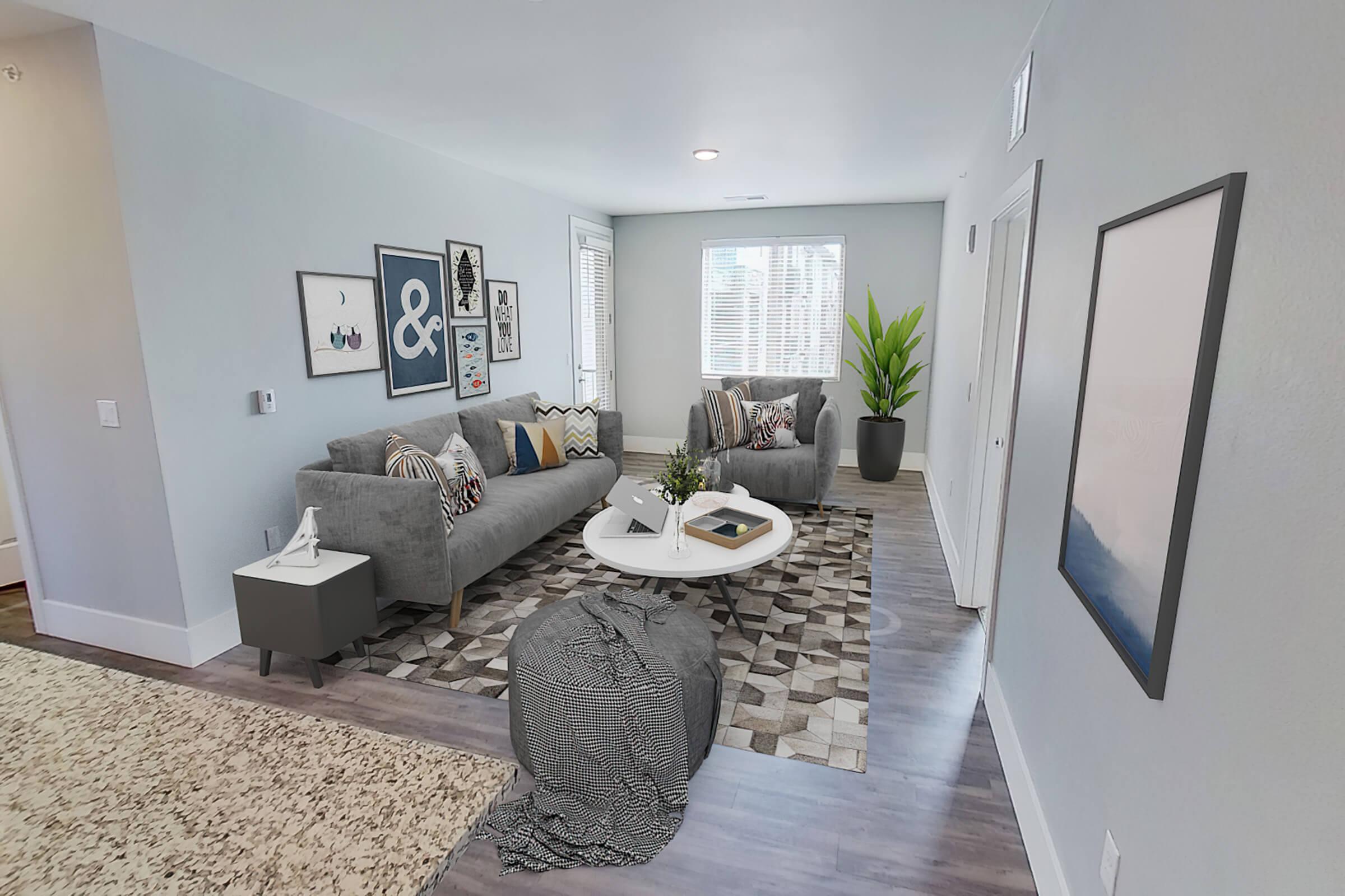 A modern living room featuring two gray sofas adorned with decorative pillows, a round coffee table, and a patterned area rug. Bright natural light enters through window blinds, highlighting framed artwork on the walls and a potted plant in the corner, creating a stylish and inviting atmosphere.