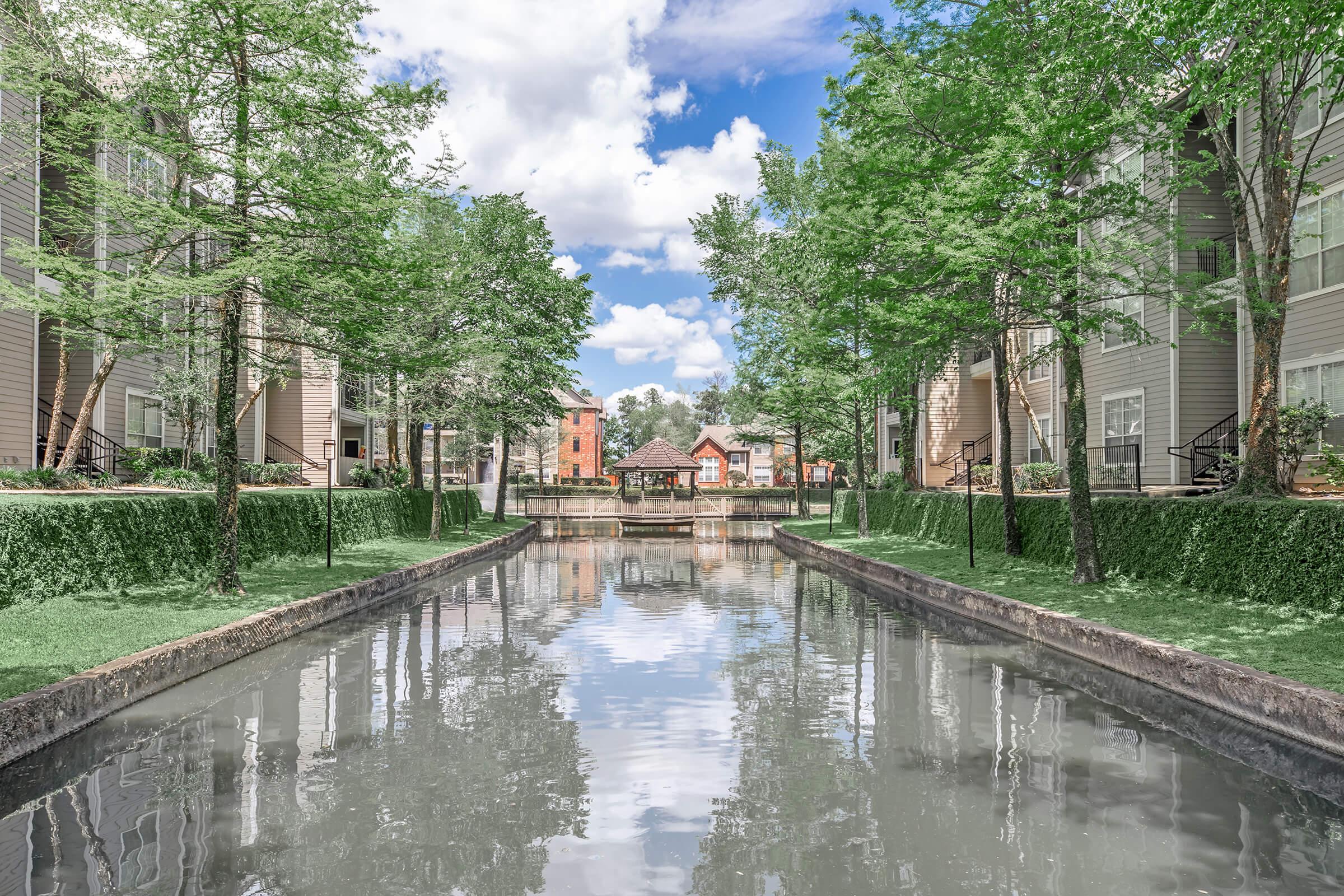 A serene view of a canal bordered by lush greenery and modern apartment buildings. The calm water reflects the clouds and surrounding trees, creating a peaceful atmosphere. In the distance, a small bridge connects pathways, enhancing the scenic charm of the community.