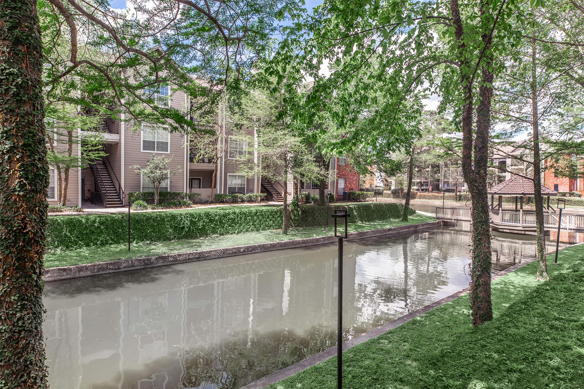 Tranquil scene of a landscaped area featuring a narrow canal surrounded by green grass and trees. In the background, there are residential apartment buildings with balconies. Soft sunlight filters through the leaves, creating a peaceful atmosphere ideal for relaxation or a leisurely walk.