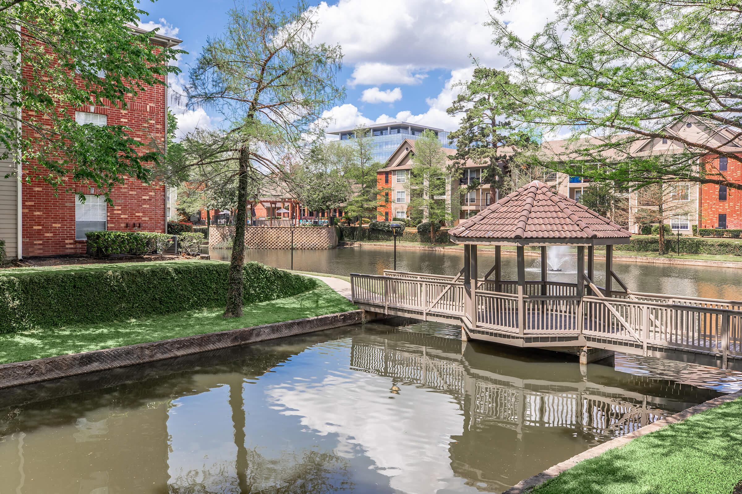 A serene view of a landscaped area with a wooden gazebo by a calm waterway. Surrounding trees reflect in the water, and residential buildings line the opposite bank under a bright blue sky with scattered clouds.