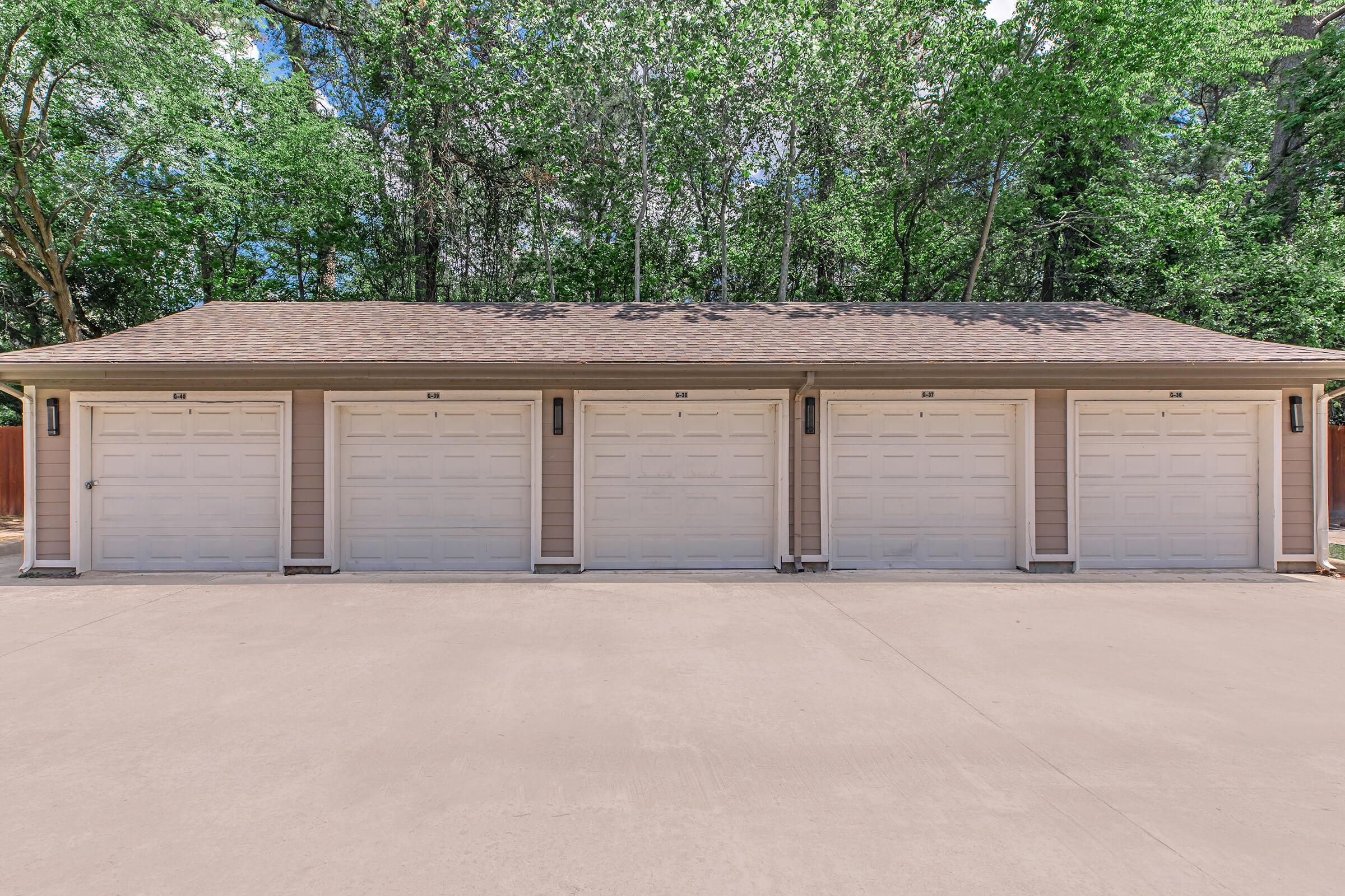 A row of four white garage doors on a single-story building, set against a backdrop of lush, green trees. The driveway is concrete, and the scene is bright and airy, suggesting a well-maintained property. The garages are uniform in style, with a simple, clean design.