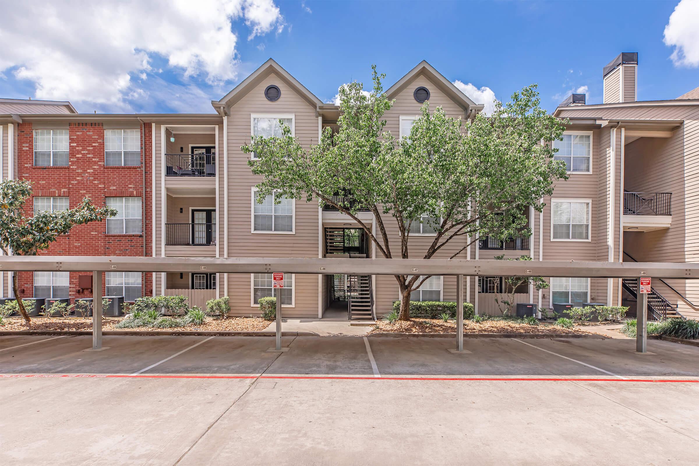 A multi-story apartment building with a mix of brick and beige siding. There are balconies on the upper floors, and a large tree in front provides some shade. The parking area is visible in the foreground, with marked spaces and a clear blue sky in the background.