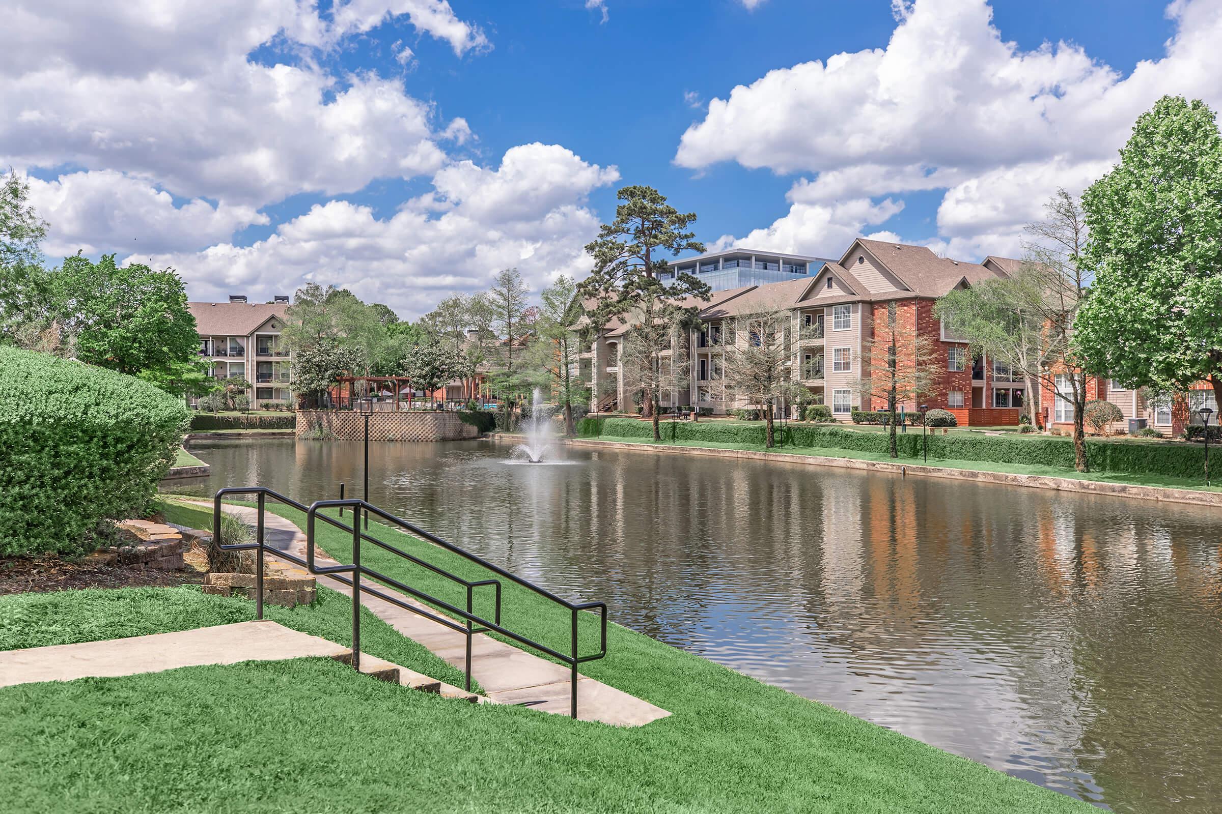 A serene lakeside view featuring a peaceful water pond surrounded by green grass and well-maintained landscaping. In the background, residential buildings blend with the natural scenery, alongside a bridge and fluffy clouds scattered across a bright blue sky.
