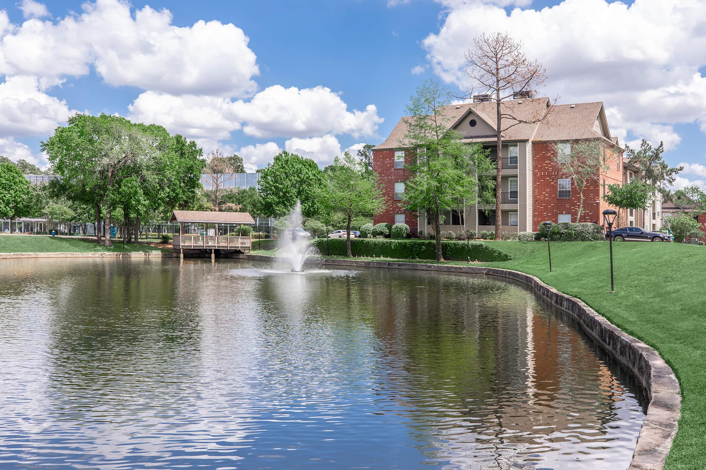 A serene pond with a fountain in the center, surrounded by lush green trees and grass. A two-story brick building is visible in the background, along with a wooden dock extending over the water. The scene is set under a bright blue sky dotted with fluffy white clouds.