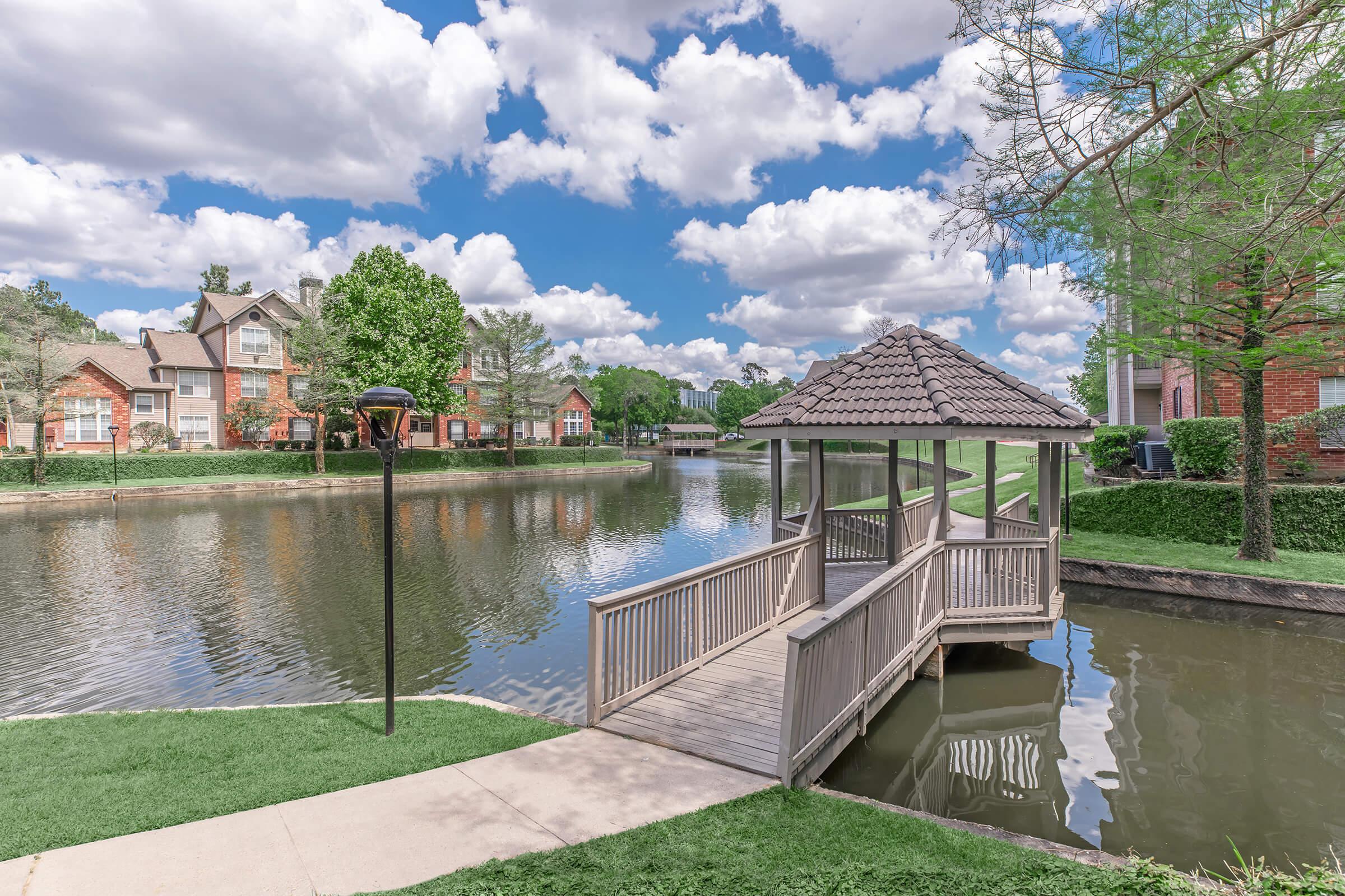 A scenic view of a pond with a wooden gazebo extending over the water. Lush greenery surrounds the pond, and residential buildings with brick facades are visible in the background. The sky is bright and partly cloudy, enhancing the tranquil atmosphere of the setting.