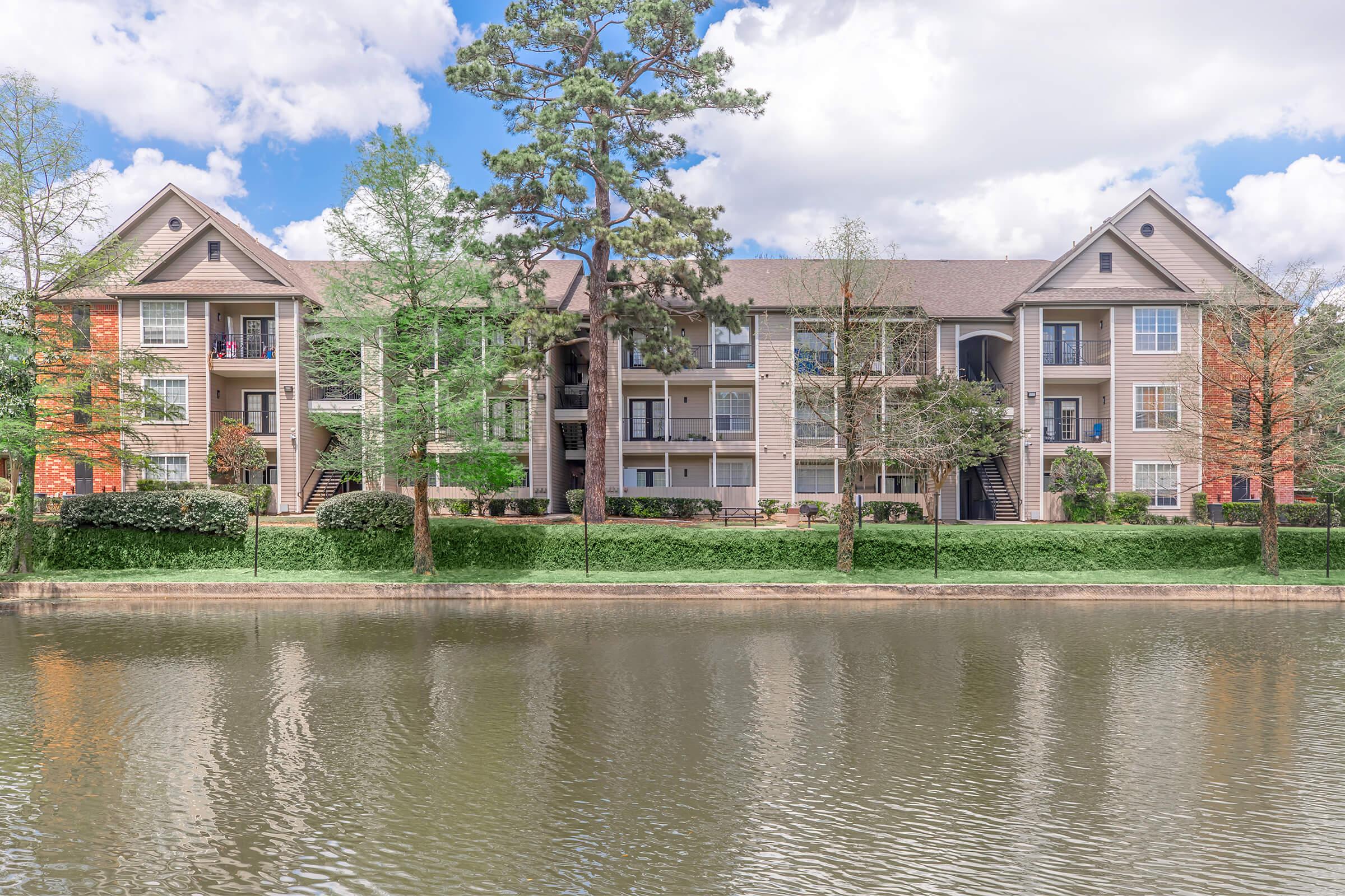 A view of a residential apartment complex by a calm lake, featuring multiple buildings with balconies, surrounded by trees and well-maintained landscaping under a partly cloudy sky. The scene reflects a peaceful and inviting atmosphere.