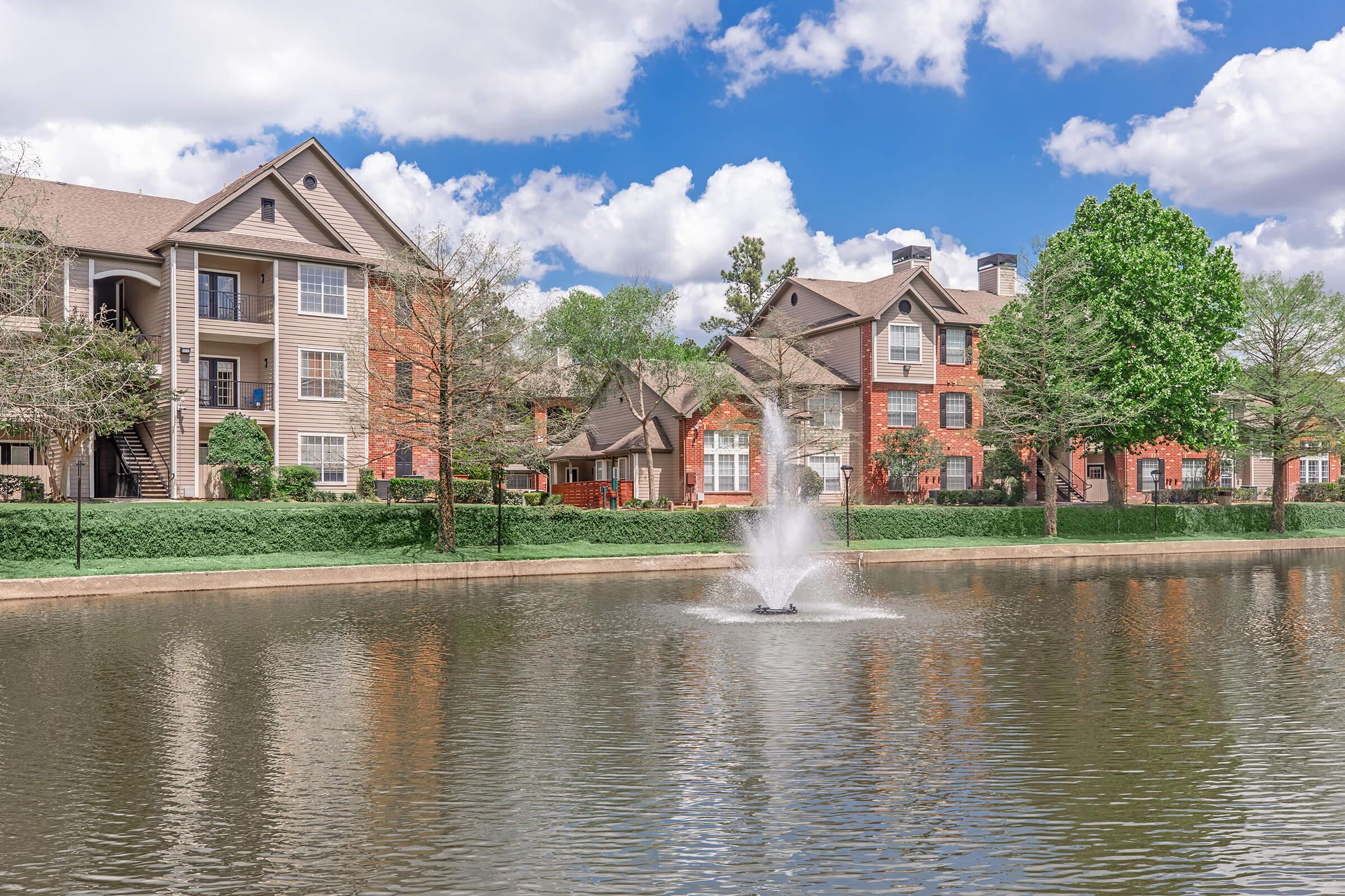 A serene lake surrounded by brick apartment buildings, with a fountain creating a gentle spray. Lush green trees line the water, and the sky is partly cloudy with blue peeking through. The scene captures a tranquil residential community setting.