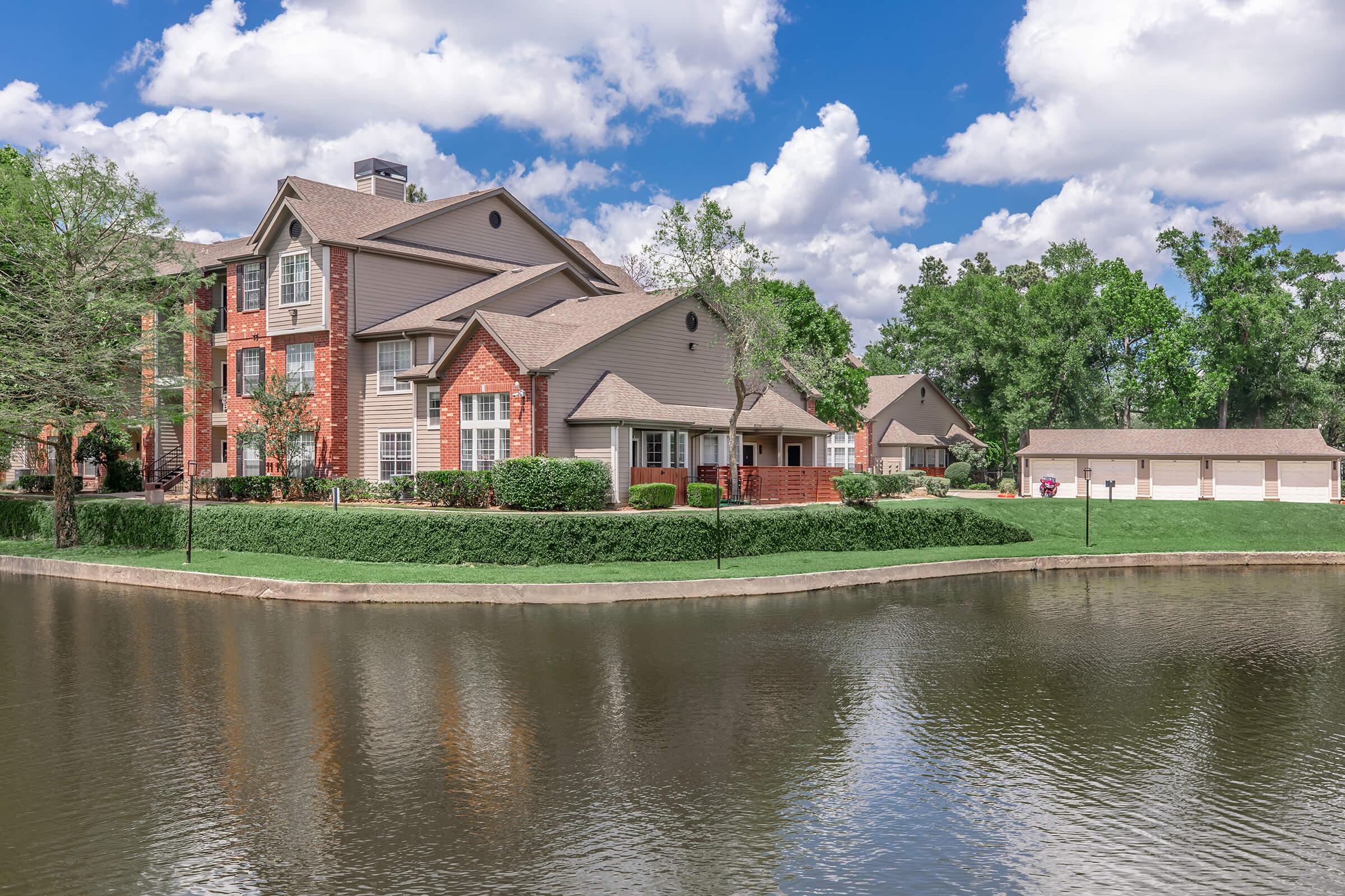 A picturesque residential complex next to a serene lake, featuring brick and light brown buildings surrounded by lush greenery. The sky is partly cloudy, adding to the tranquil atmosphere of the scene.