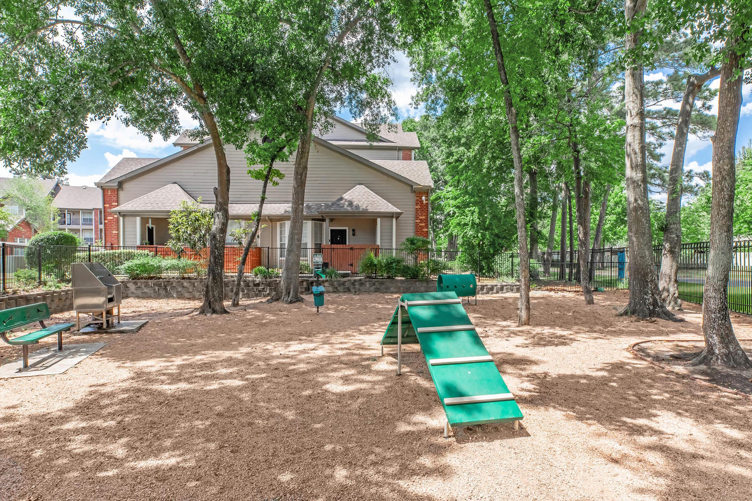 A playground area featuring a green slide, climbing structure, and fenced yard. The ground is covered with wood chips, surrounded by trees. In the background, a large house with a sloped roof is visible, along with a picnic table and grill. A sunny sky adds brightness to the scene.