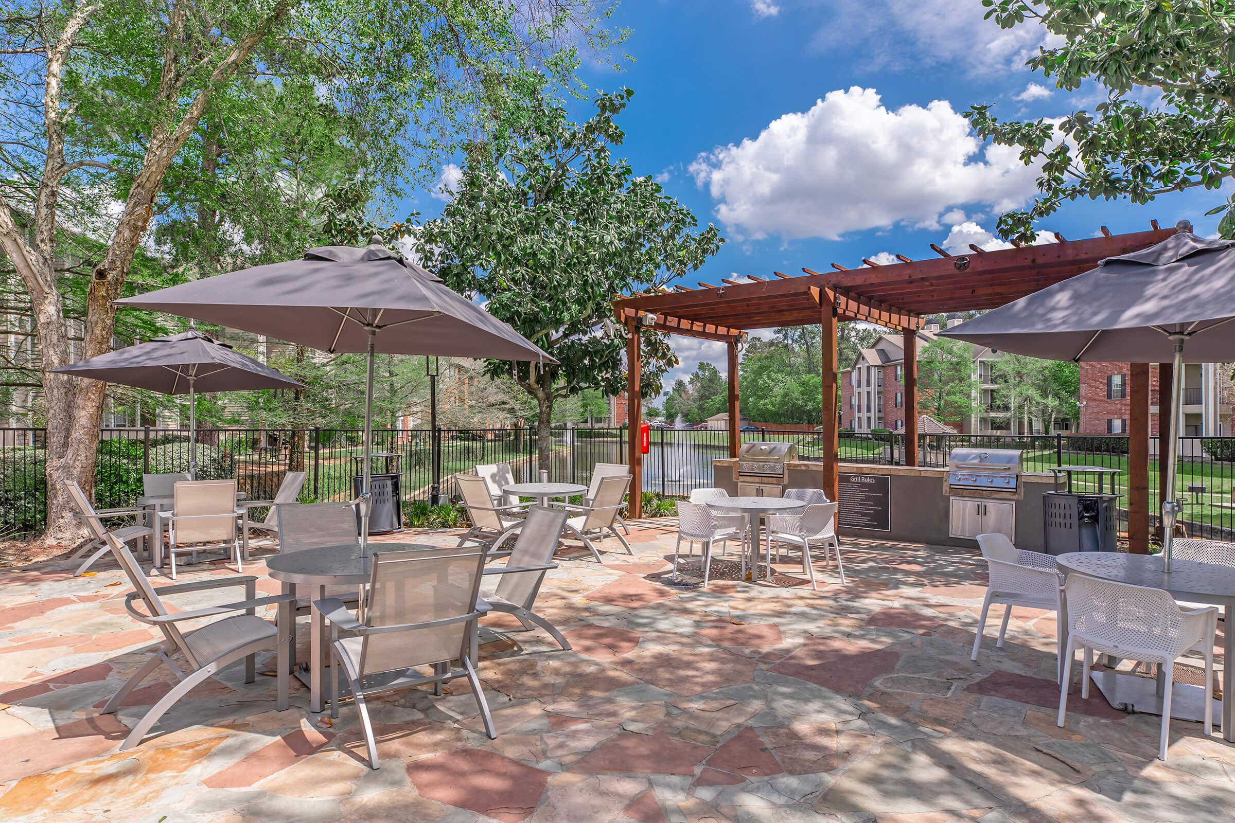 Outdoor seating area featuring several tables and chairs under gray umbrellas. There's a wooden pergola with a grill and a stone pathway. Lush green trees surround the space, and a well-maintained lawn is visible in the background under a clear blue sky with some fluffy white clouds.
