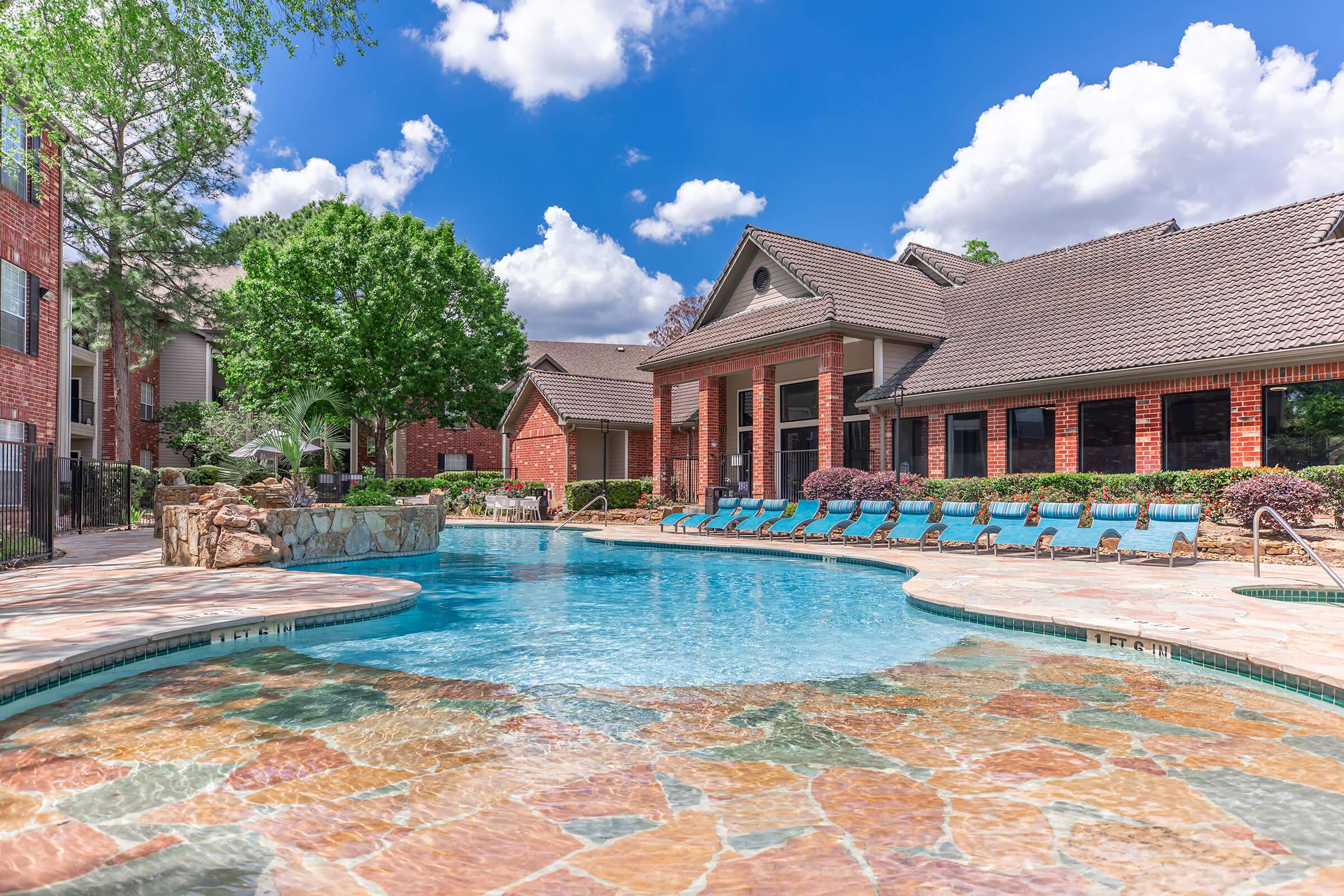 A clear blue swimming pool surrounded by natural stone and landscaped grass, with lounge chairs lined up nearby. In the background, a brick building with large windows and a well-maintained garden area. Bright blue skies with fluffy white clouds add to the inviting atmosphere.