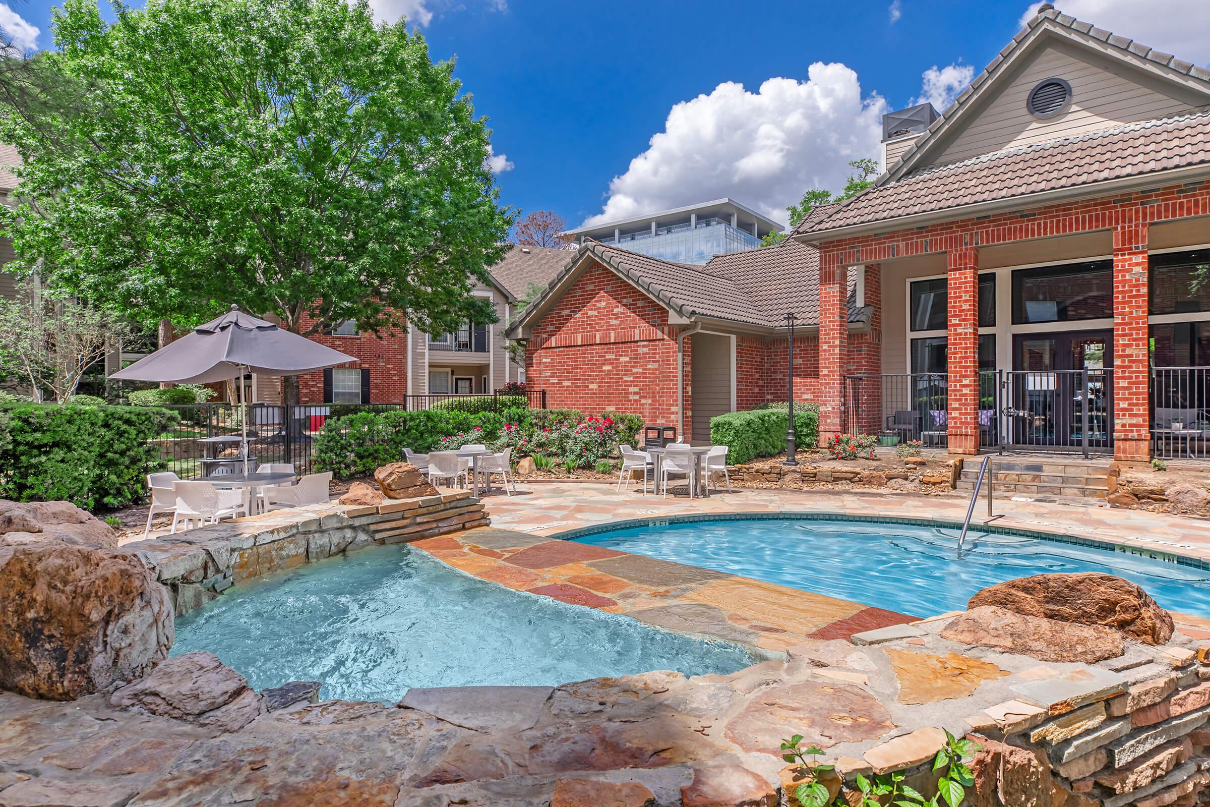 A tranquil outdoor pool area featuring a stone-bordered pool with a small waterfall, surrounded by lush greenery and patio seating. Bright blue skies with fluffy clouds enhance the inviting atmosphere of the residential complex.