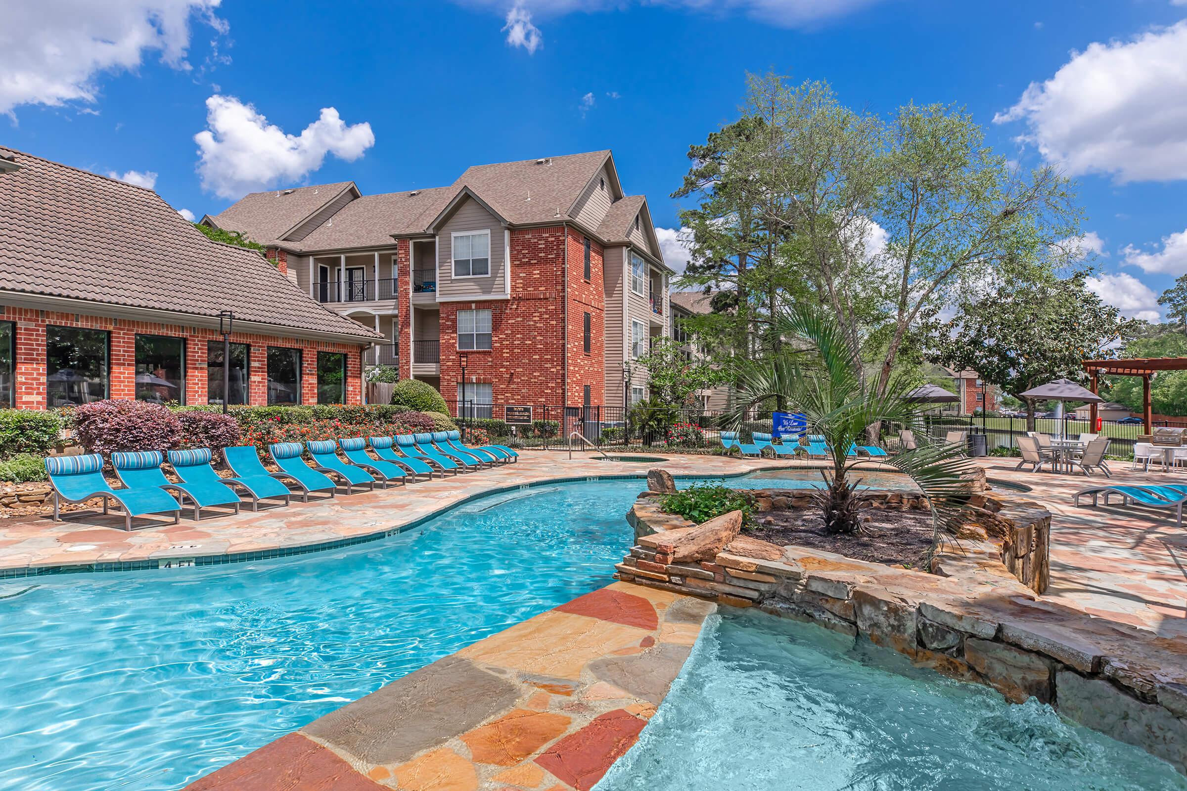 A serene outdoor pool area featuring a curved swimming pool surrounded by lounge chairs. In the background, there are residential buildings with brick and stucco exteriors, complemented by lush landscaping and blue skies with fluffy white clouds. An inviting space for relaxation and leisure.