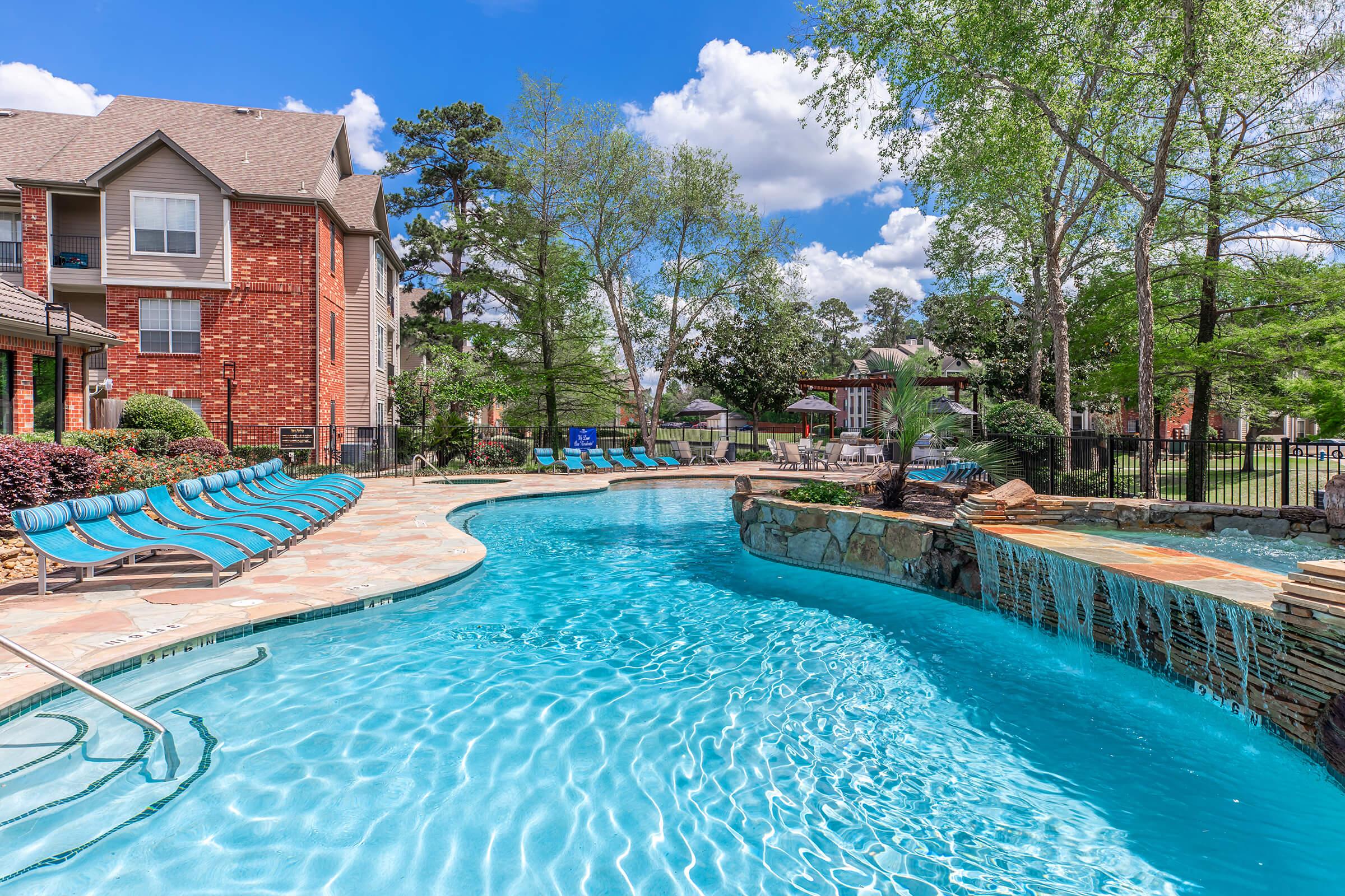 A serene outdoor pool area featuring a curvy pool with clear blue water. Lounge chairs line the poolside, surrounded by trees and greenery. In the background, a brick apartment building can be seen, along with a waterfall feature and a fenced area, all under a bright blue sky with fluffy clouds.