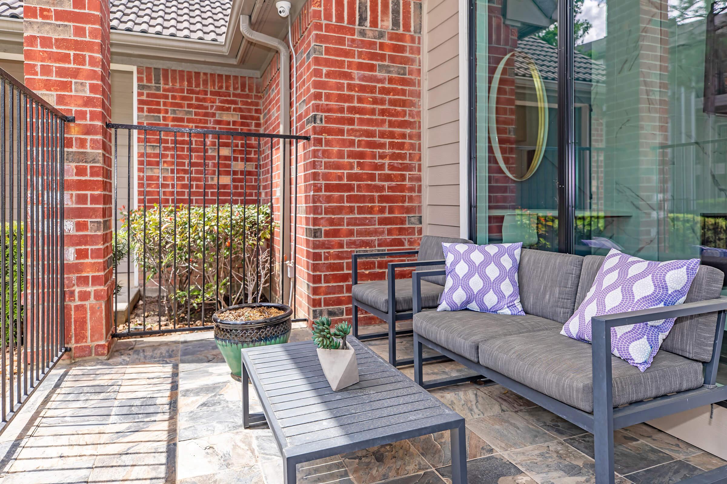 A cozy outdoor patio featuring a gray sofa with purple patterned pillows, accompanied by a small black coffee table. The area is surrounded by brick walls and green shrubs, with sunlight casting shadows on the stone floor. A potted plant sits atop the table, enhancing the inviting atmosphere.