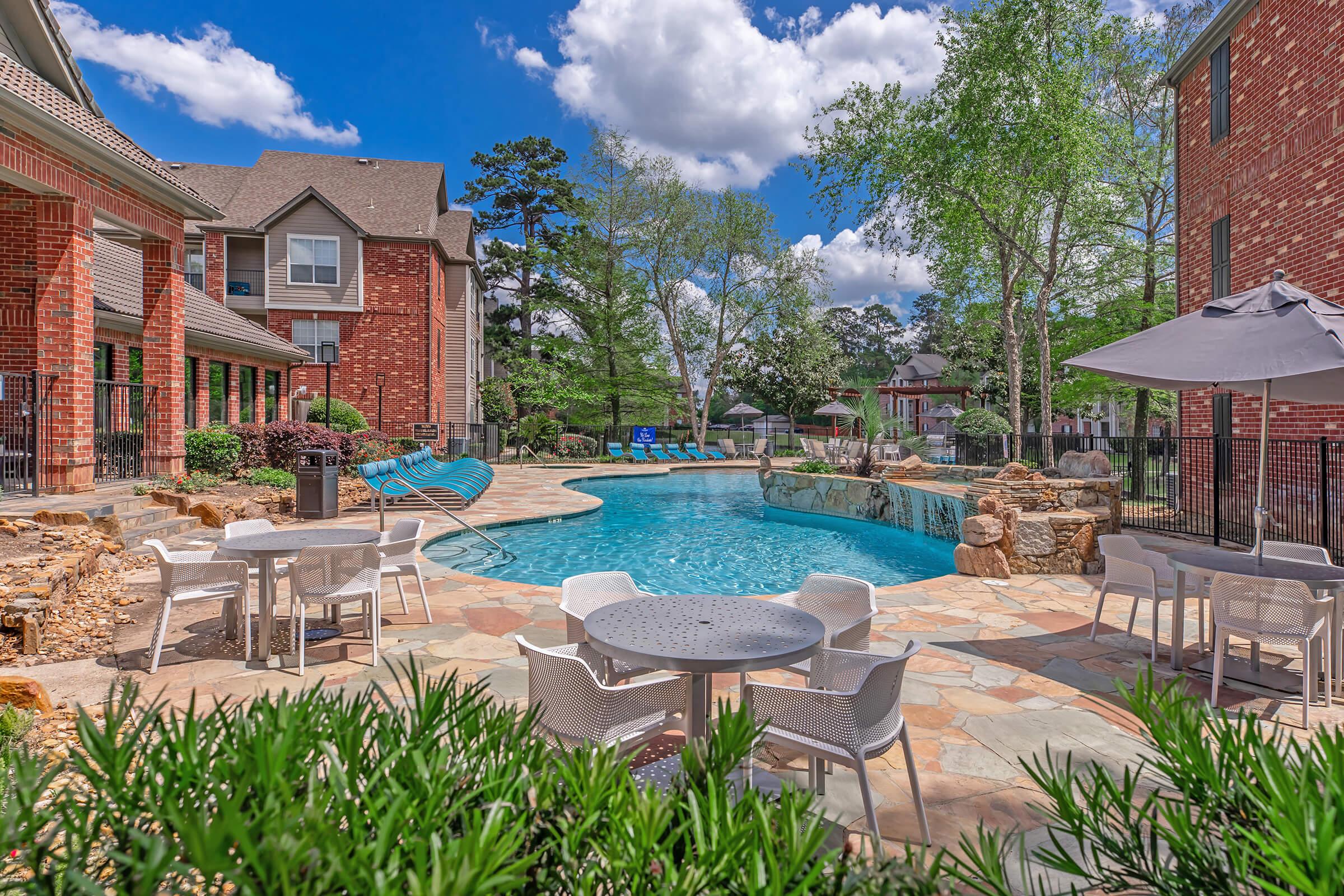 A vibrant outdoor pool area surrounded by lush greenery. The pool is inviting with clear blue water, and there are several white lounge chairs and tables set up nearby. Bright sunshine and fluffy clouds create a cheerful atmosphere, while brick buildings frame the scene in the background.