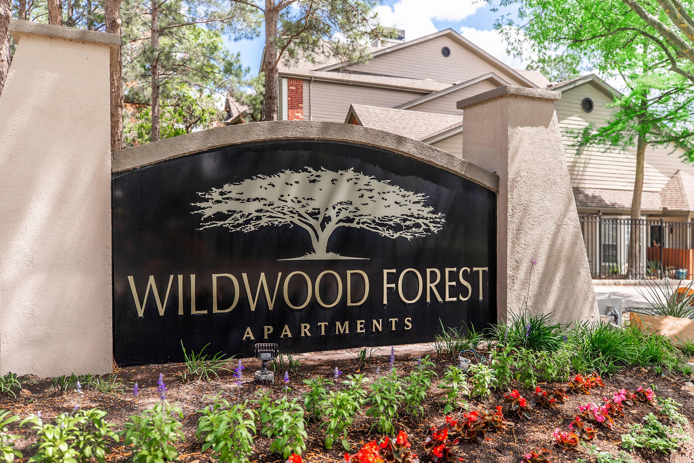 A sign for Wildwood Forest Apartments featuring a large tree graphic and surrounded by colorful flower beds. The background includes residential buildings and greenery, creating a welcoming atmosphere.