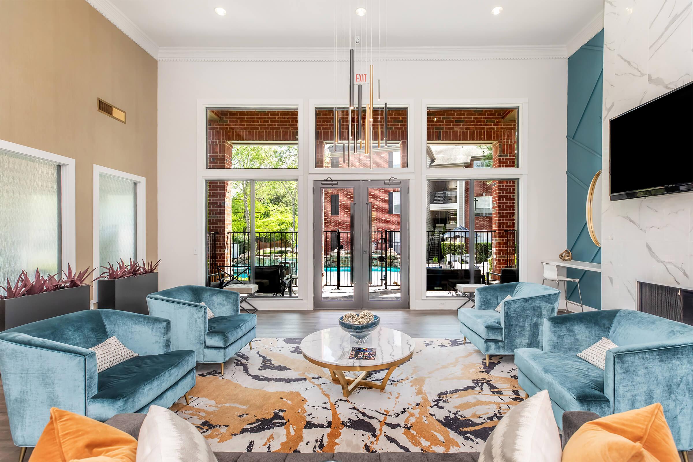 A modern lounge area featuring two teal velvet armchairs, a decorative coffee table with a bowl, and an area rug in warm tones. Large windows provide a view of a pool area outside, while the walls are accented with neutral colors and a wall-mounted TV adds a contemporary touch.