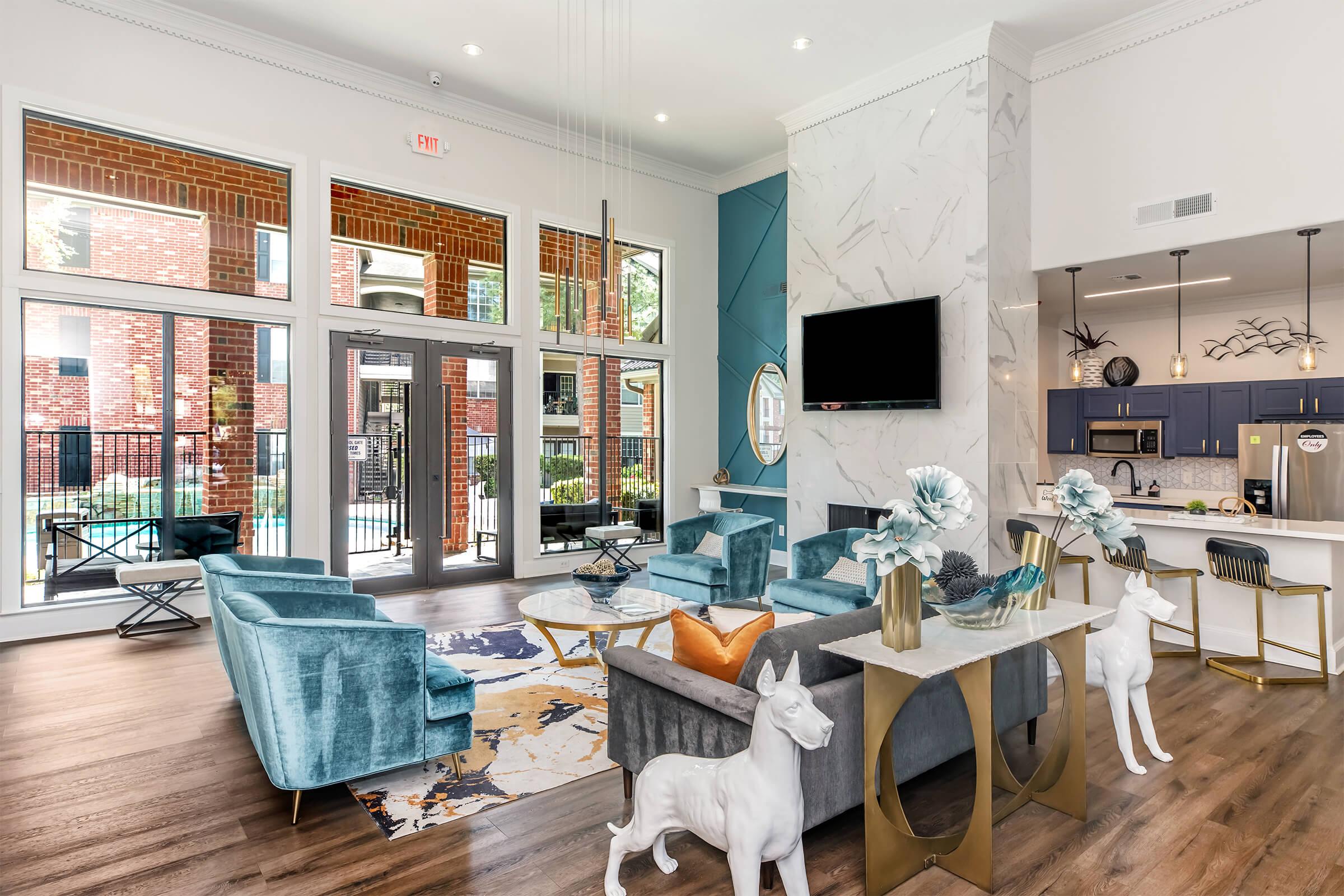 A modern lounge area featuring plush turquoise sofas, a glass coffee table, and decorative sculptures. Large windows allow natural light, showcasing a view of the outdoors. There's a contemporary kitchen in the background with stainless steel appliances and stylish accents, creating a sleek and inviting atmosphere.
