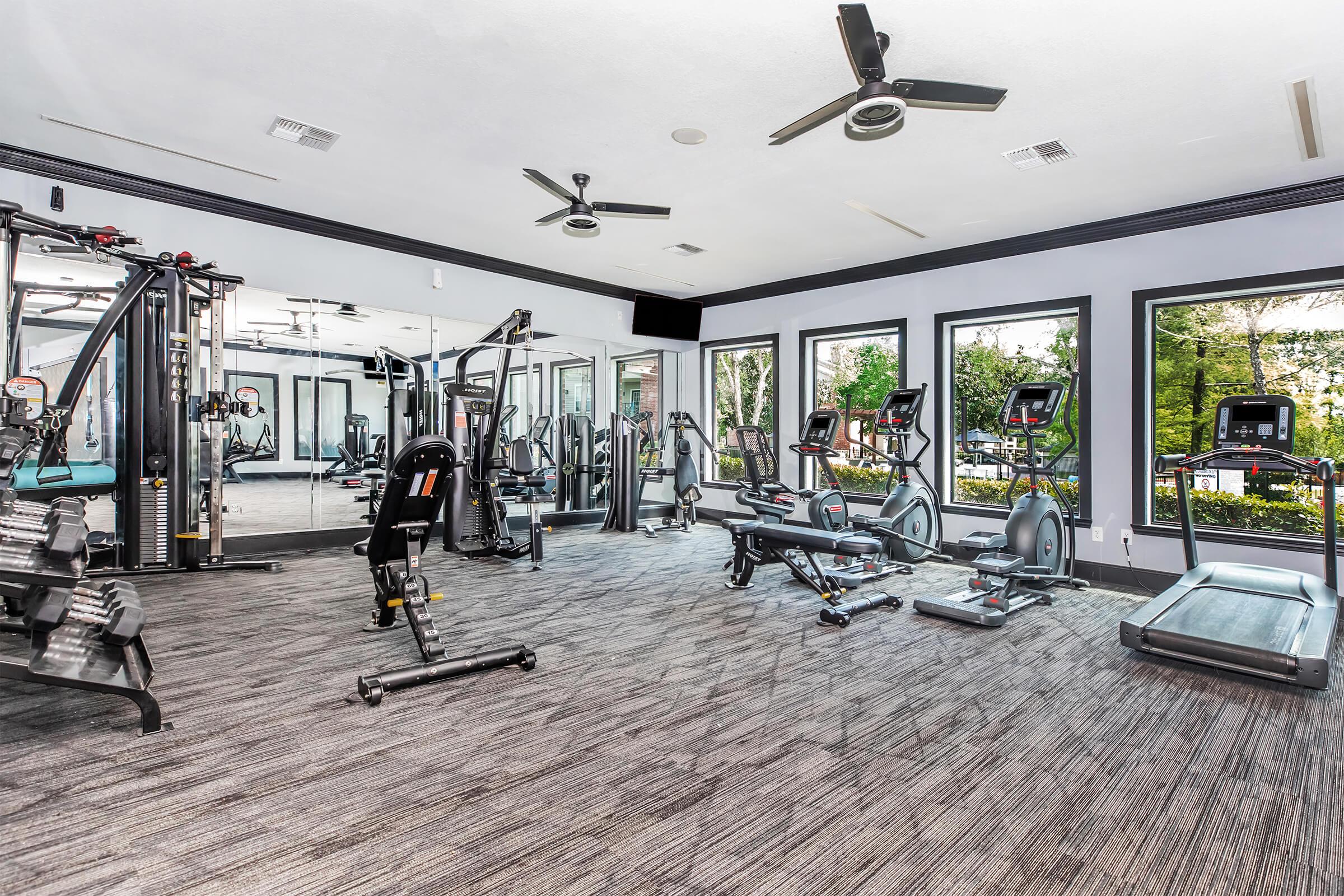 A modern gym interior featuring various exercise equipment, including treadmills, stationary bikes, weight machines, and free weights. The space is well-lit with large windows allowing natural light, and has a clean, open layout with a neutral color scheme and ceiling fans for ventilation.
