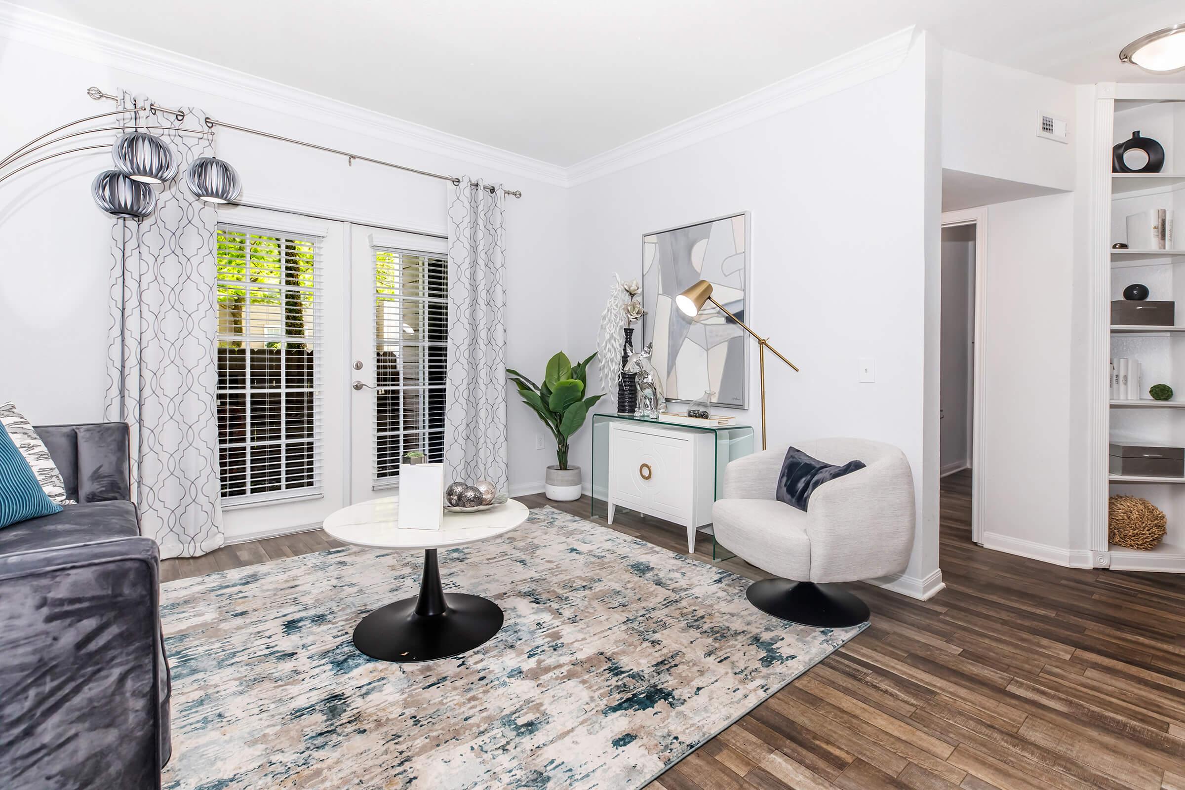 A modern living room featuring a gray sofa and a round marble coffee table on a patterned area rug. There are light-colored curtains, potted plants, and a decorative console table with a framed mirror. The room has hardwood flooring and bright natural light coming through the windows.