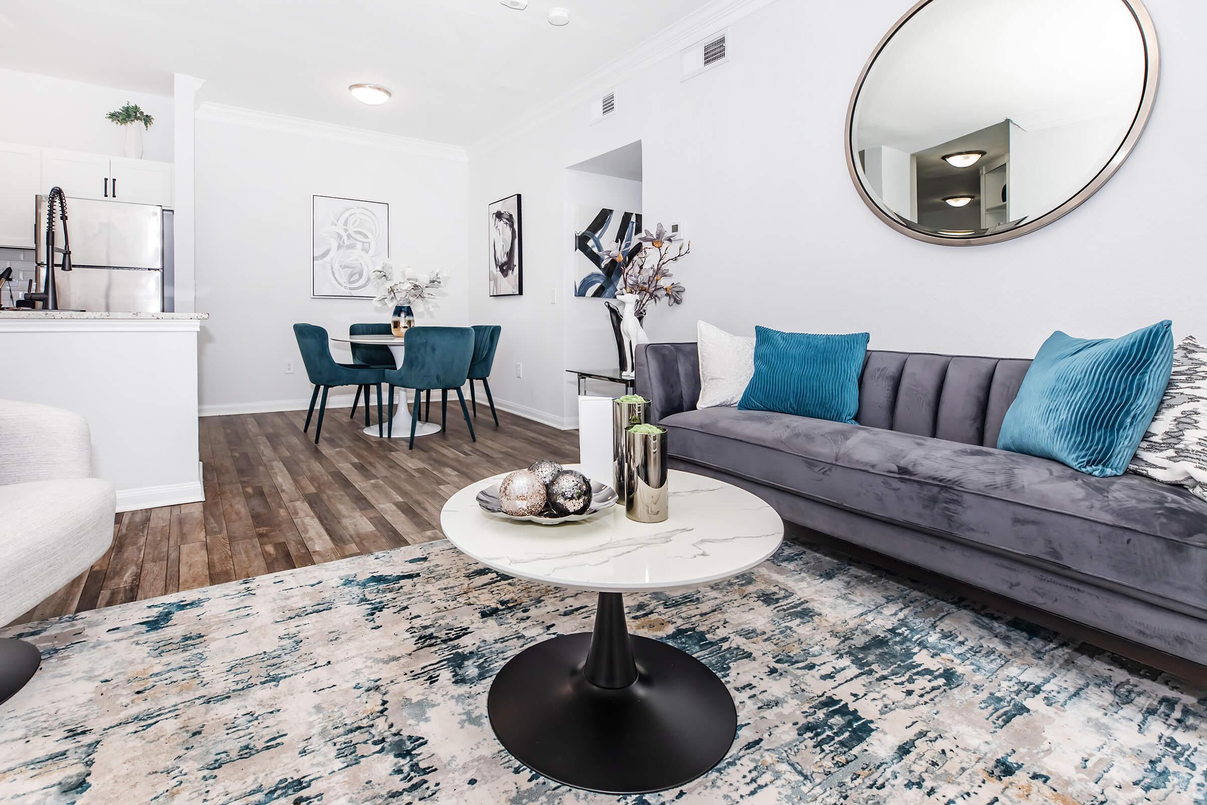 A modern living room featuring a plush gray sofa with blue and white cushions, a round coffee table on a decorative rug, and a dining area in the background. A large mirror adorns the wall, reflecting natural light and creating a spacious atmosphere. The decor is stylish and contemporary.