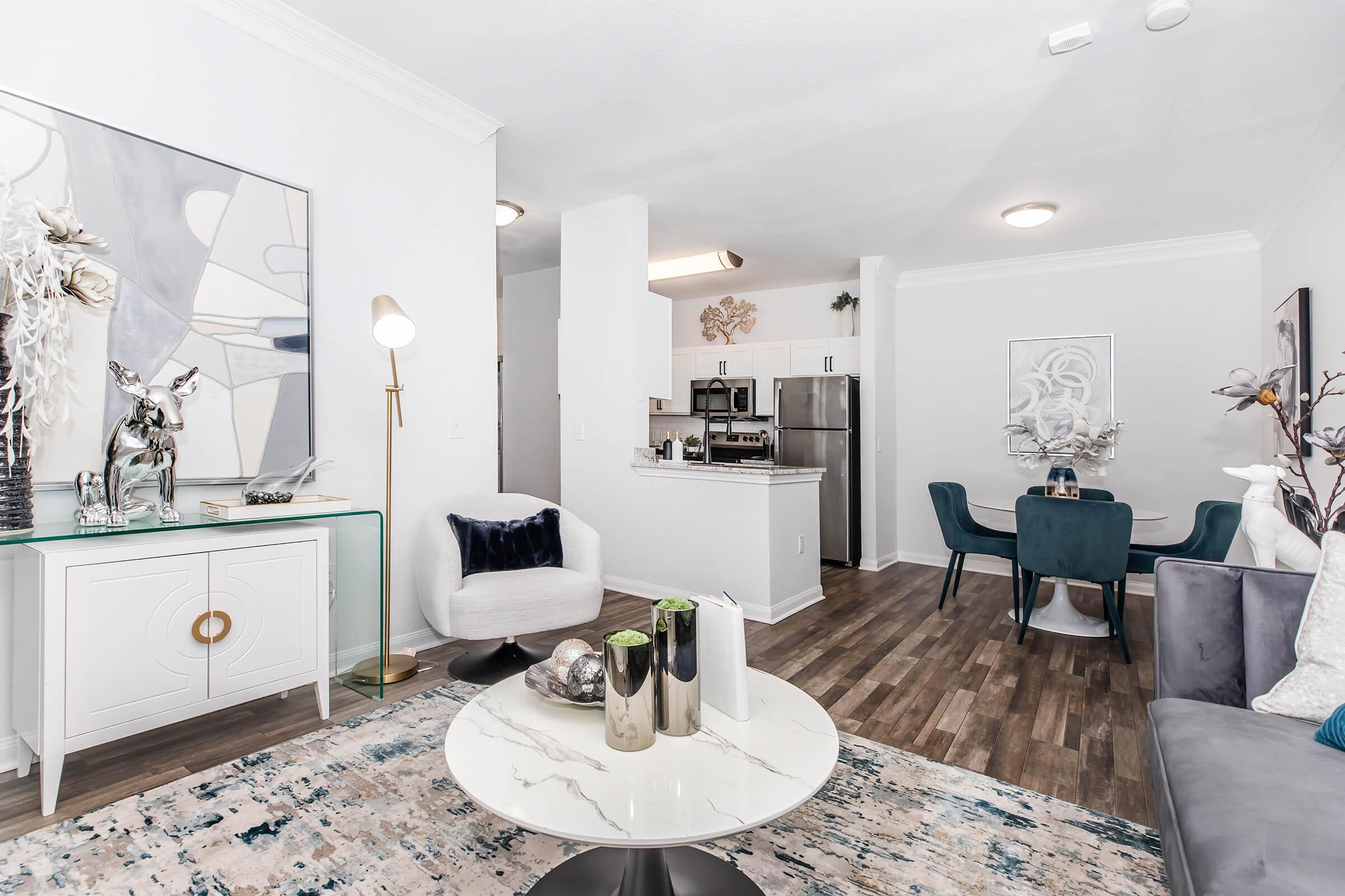 A modern living room featuring a gray sofa, a round marble coffee table, and a decorative console table with sculptures. The space includes a dining area with a round table and teal chairs. The walls are adorned with abstract art, and large windows allow natural light to brighten the room.