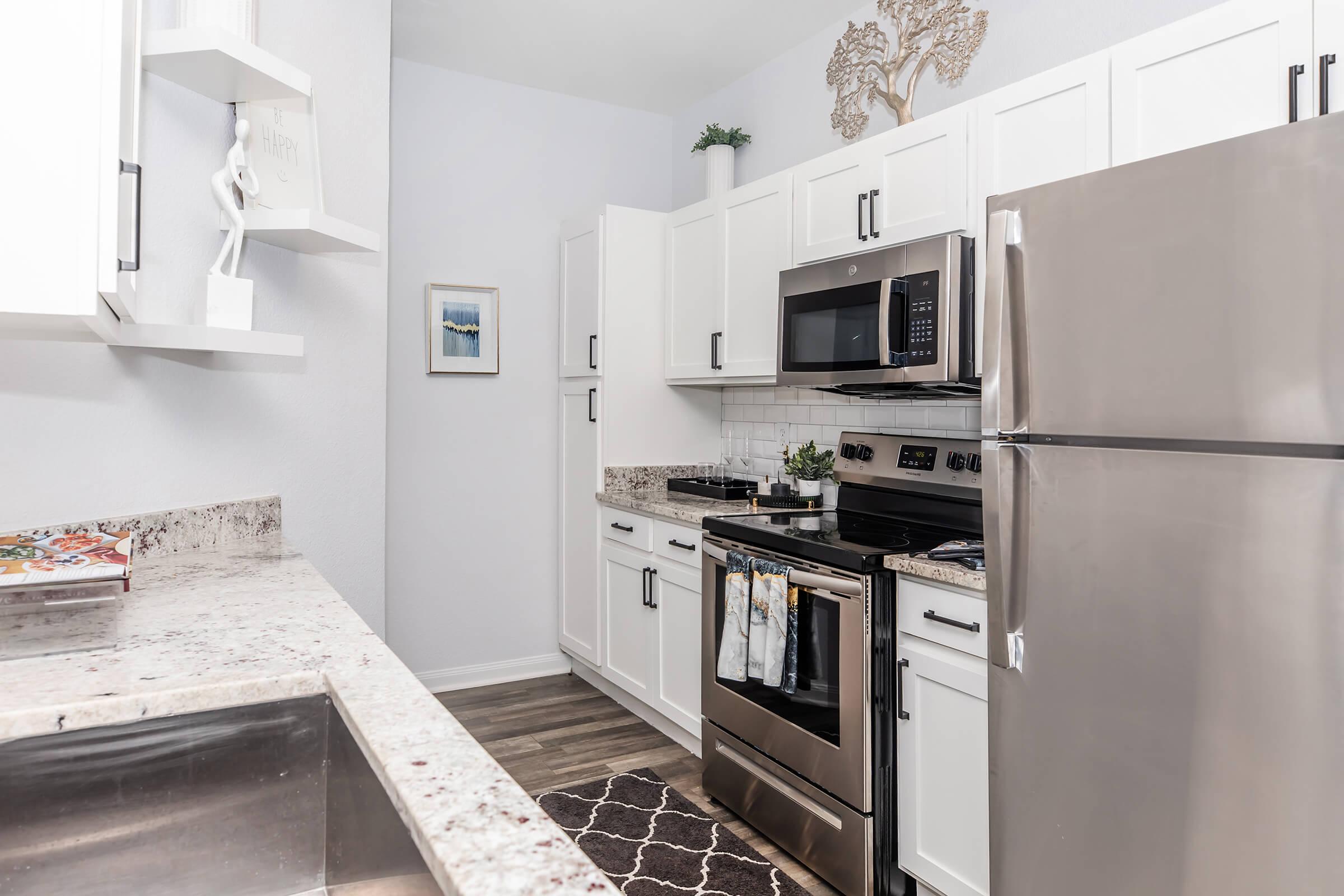 A modern kitchen featuring white cabinetry, stainless steel appliances, and a granite countertop. The kitchen includes an oven, microwave, and refrigerator, with a decorative wall piece and a small plant on the shelves. A dark rug adds contrast to the light-colored floor and walls.
