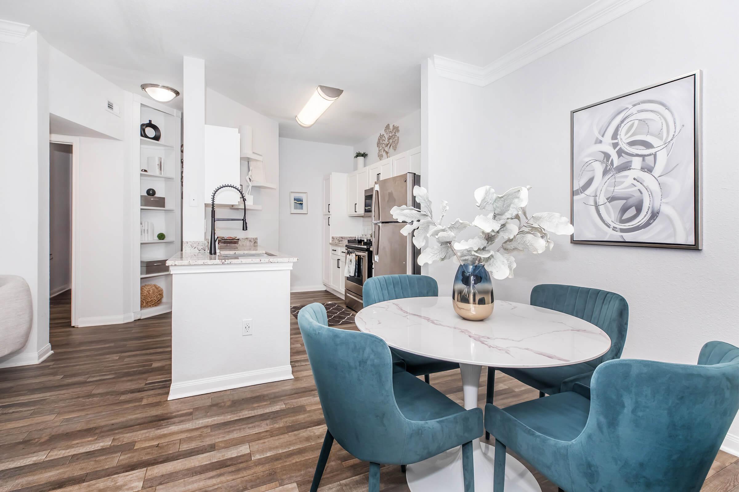 A modern kitchen and dining area featuring a round marble table with blue velvet chairs. The kitchen showcases white cabinets, stainless steel appliances, and decorative elements. A wall art piece is displayed nearby, adding a touch of elegance to the space.