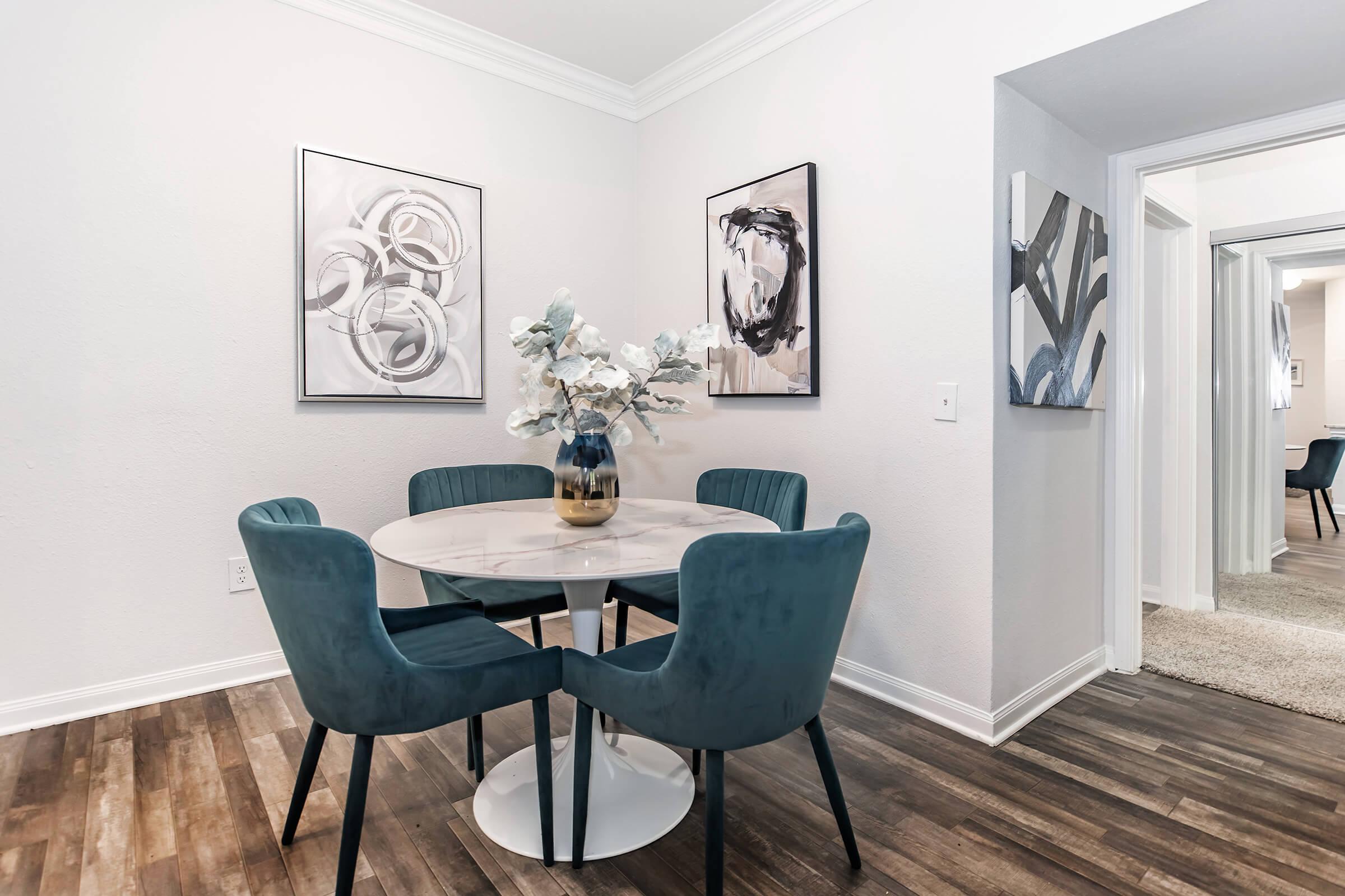 A modern dining area featuring a round marble table surrounded by four teal upholstered chairs. The walls are adorned with abstract artwork, and a vase with artificial flowers is centerpieces on the table. Light hardwood flooring complements the neutral color scheme. A doorway leads to another room in the background.