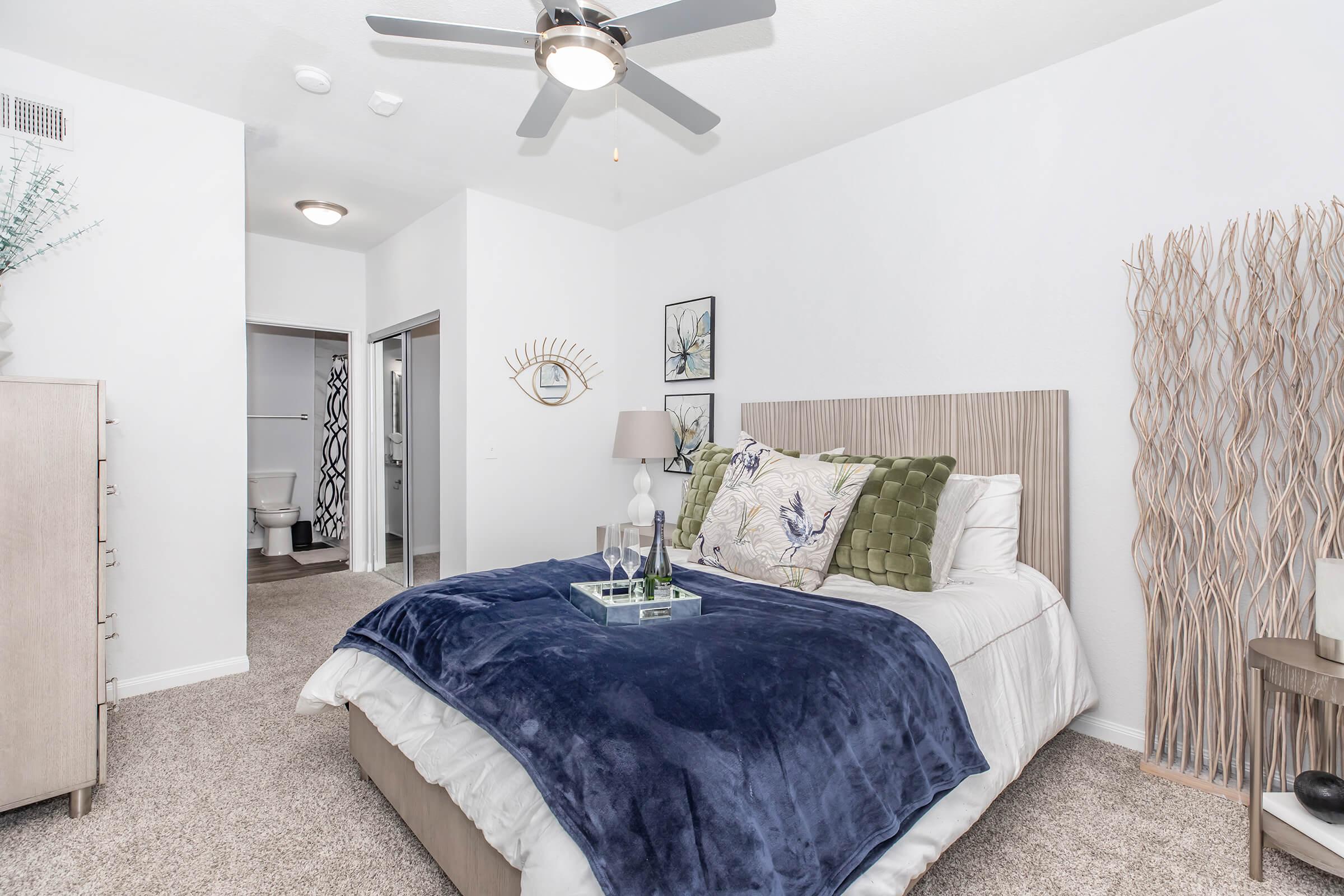 A stylish bedroom featuring a bed with a plush blue blanket and decorative pillows. There’s a small tray with items on the bed. The room has a ceiling fan, a dresser, and a textured wall accent. A doorway leads to a bathroom, and the decor includes plants and artwork, creating a cozy atmosphere.