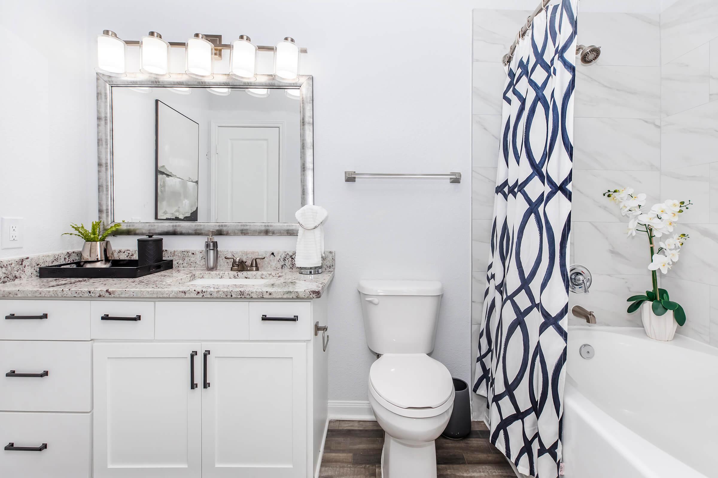 A modern bathroom featuring a white vanity with a marble countertop, a large mirror with five light fixtures above, and a decorative plant. There is a white toilet and a bathtub with a shower curtain displaying a blue and white geometric pattern. The floor is tiled in dark wood.