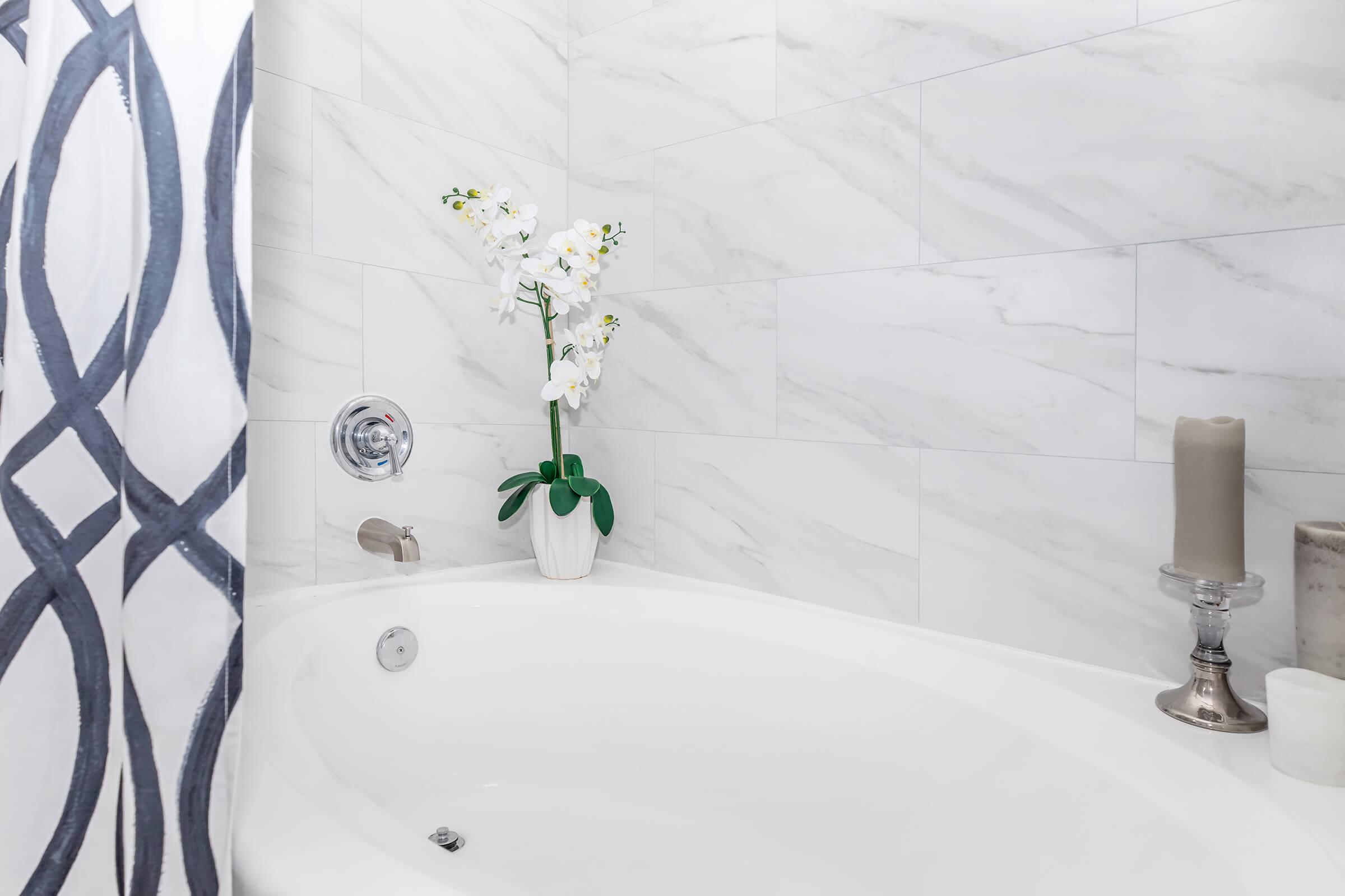 A modern bathroom featuring a white bathtub, marble tiled walls, and a decorative arrangement of orchids in a vase. A candle holder with candles is visible nearby, alongside a patterned shower curtain. The overall design is sleek and contemporary, creating a relaxing atmosphere.