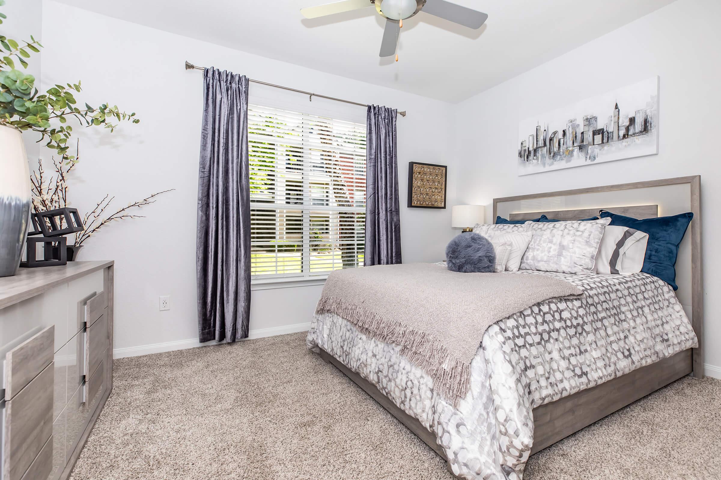 A cozy bedroom featuring a large bed with patterned bedding and accent pillows, a decorative throw, and a plush round pillow. There are elegant curtains over the window, allowing natural light to enter. A modern dresser and wall art complement the soft color palette and relaxed ambiance of the room.