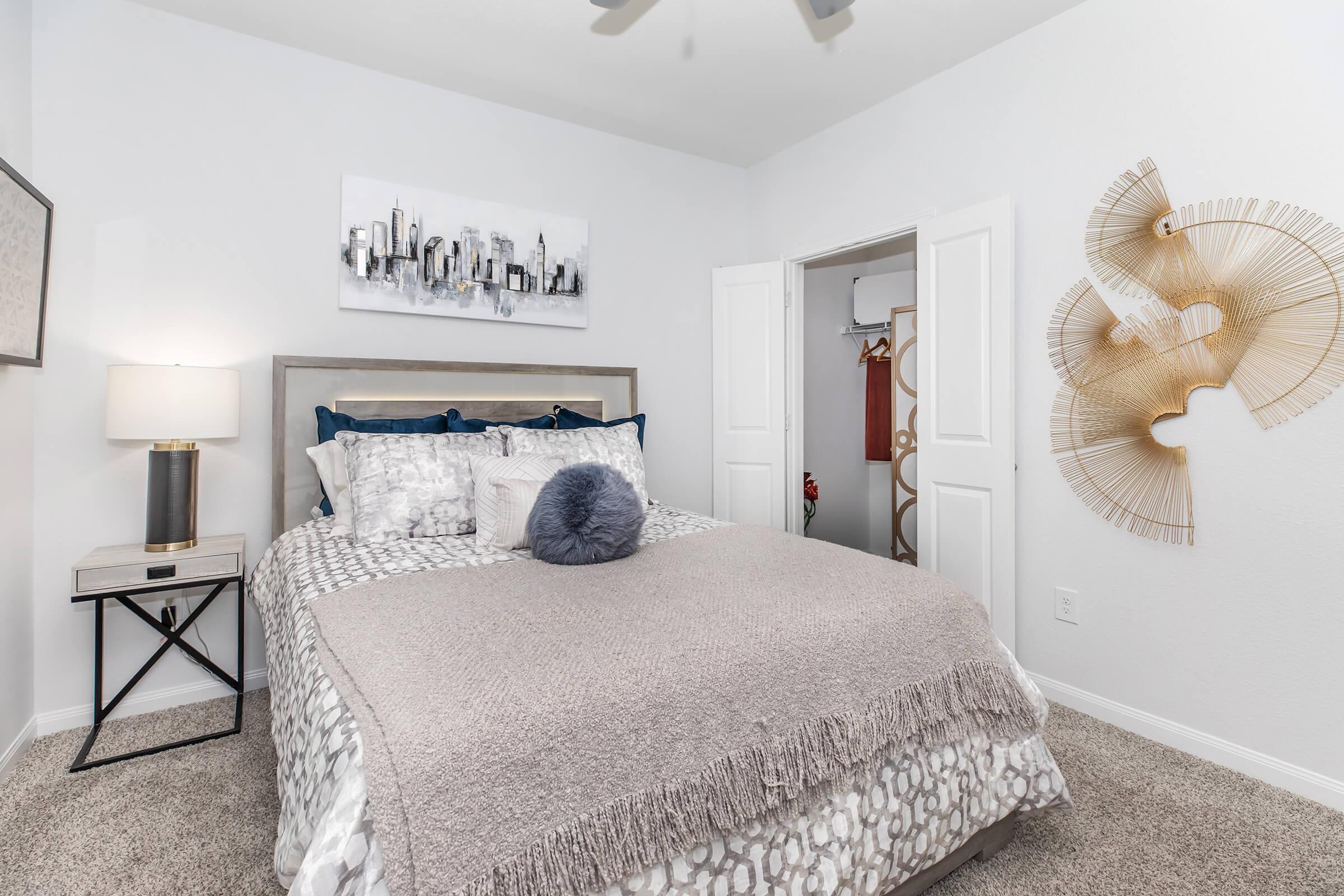Cozy bedroom featuring a comfortable bed with decorative pillows and a soft throw blanket. The room has light-colored walls, a stylish lamp on a nightstand, and a decorative wall art piece. A closet with sliding doors is visible, and the flooring is carpeted for warmth.