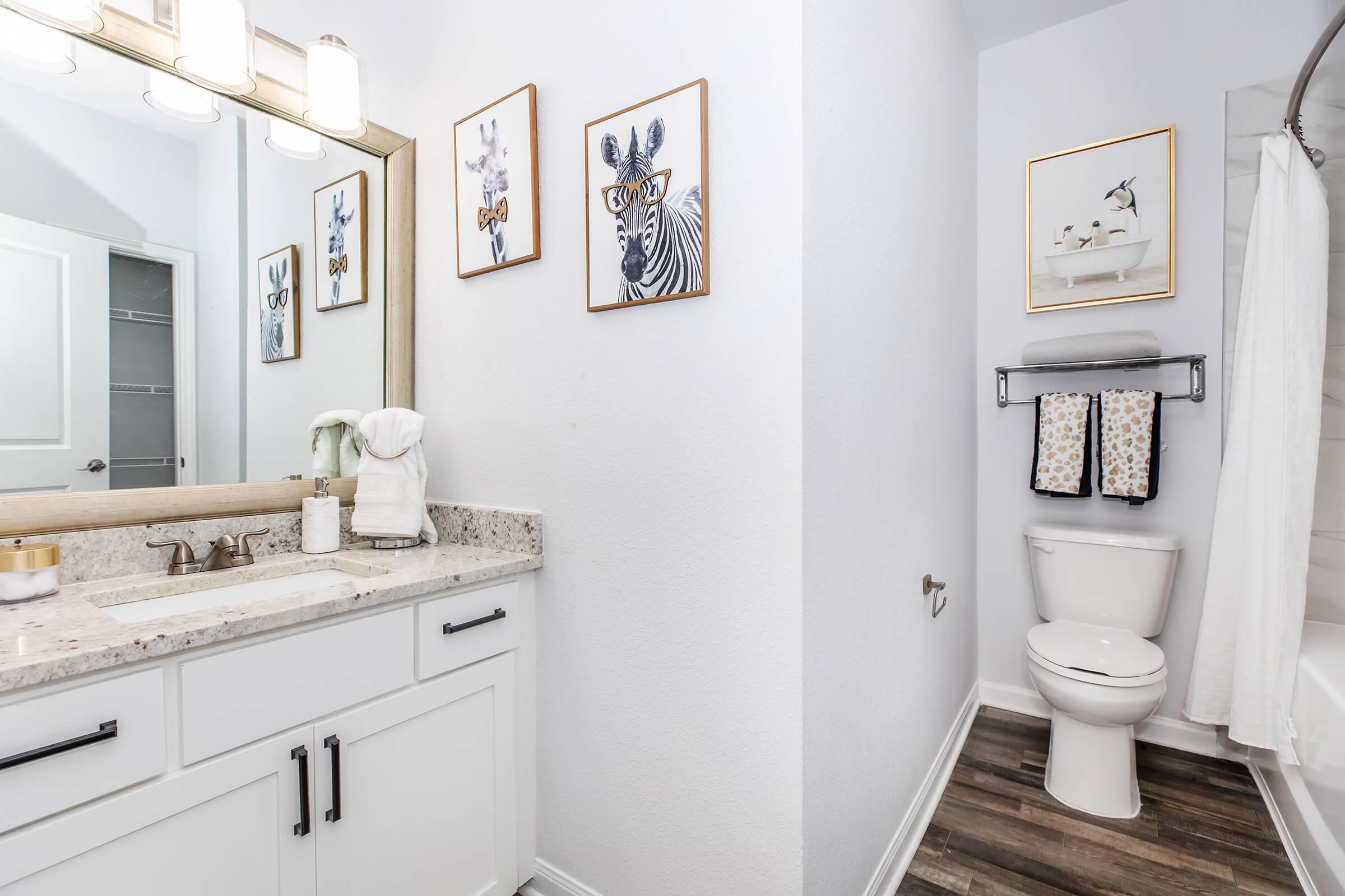 Bright and modern bathroom featuring a double sink vanity with granite countertops. Two framed animal art pieces adorn the wall, alongside a towel rack holding patterned towels. A toilet is positioned beside a bathtub with a shower curtain. The space is well-lit and has a clean, contemporary design.