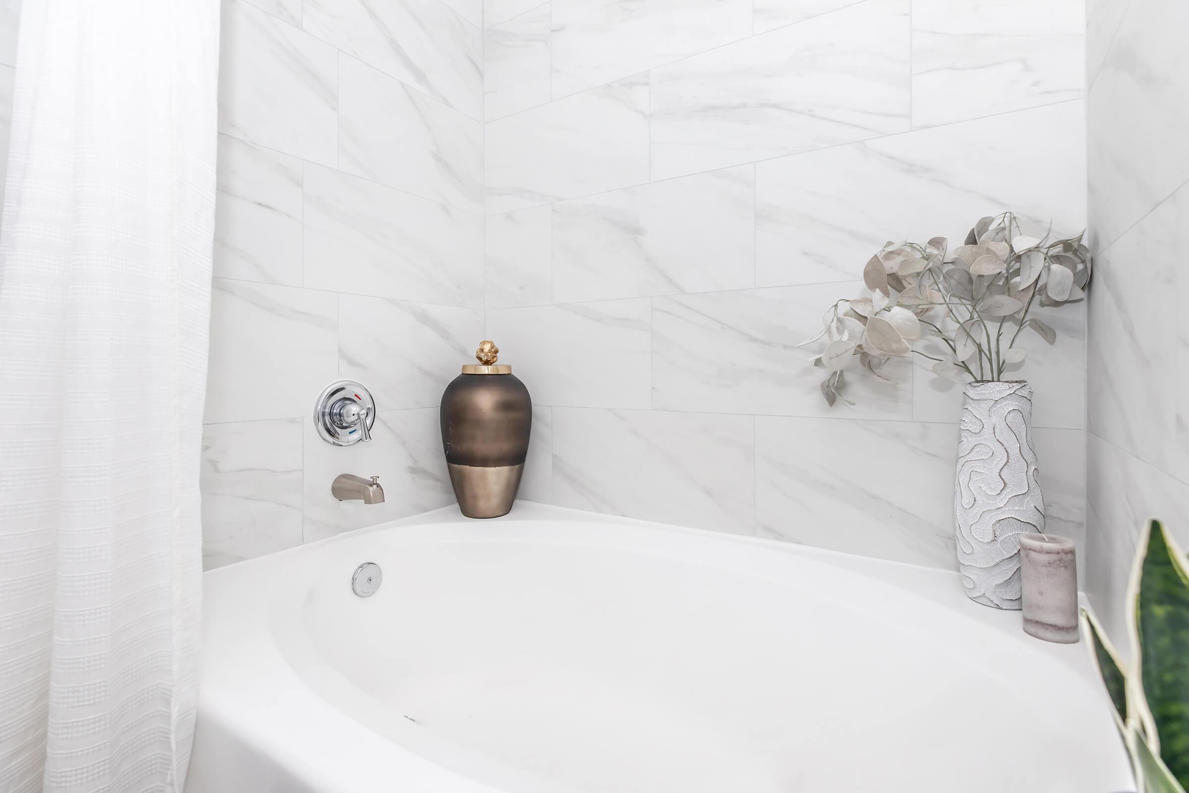 A modern bathroom featuring a white oval bathtub against a marble-tiled wall. There's a decorative grey vase with dried foliage and a sleek brown vase on the edge, creating a serene and stylish atmosphere. Soft natural light illuminates the space, enhancing the elegant design.